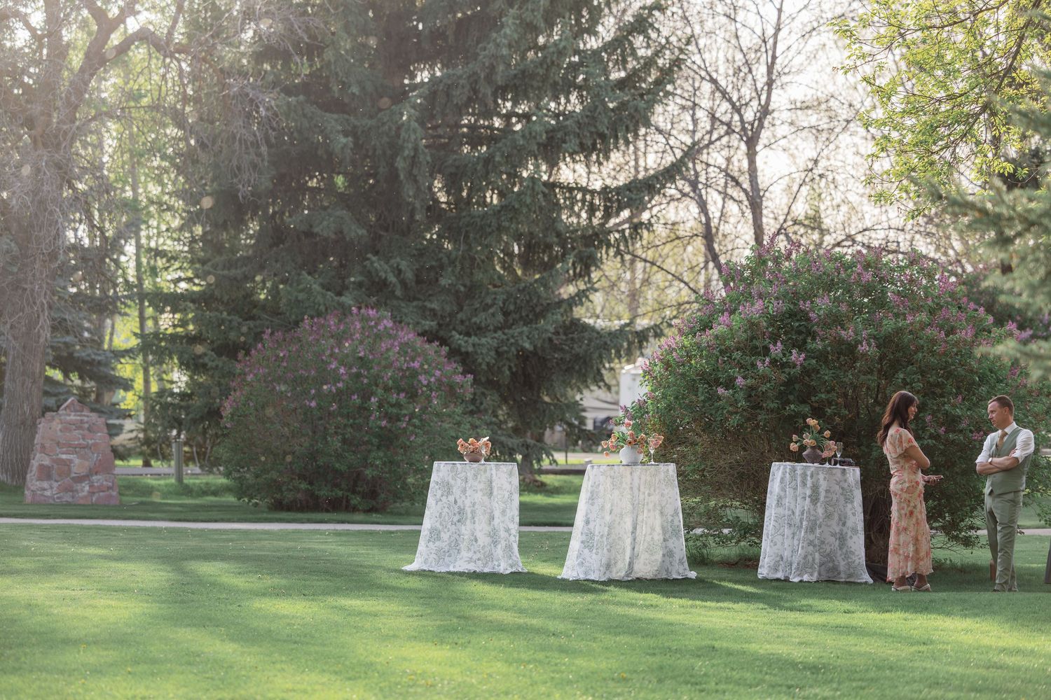Elegant outdoor wedding cocktail hour with lace-draped high tables on lush green lawn surrounded by trees.