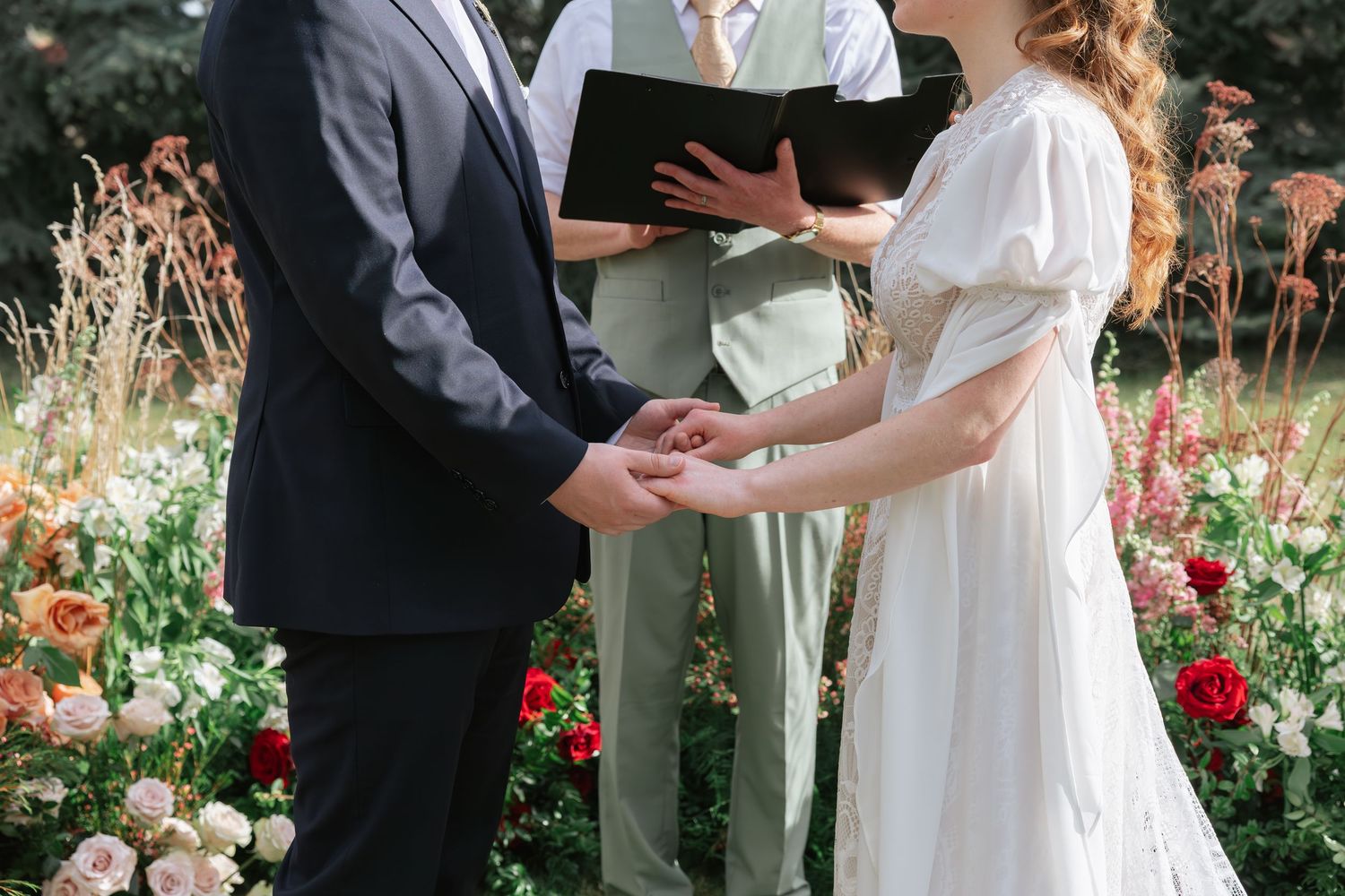 Bride and groom exchange vows holding hands during an outdoor garden wedding ceremony surrounded by colorful blooms.