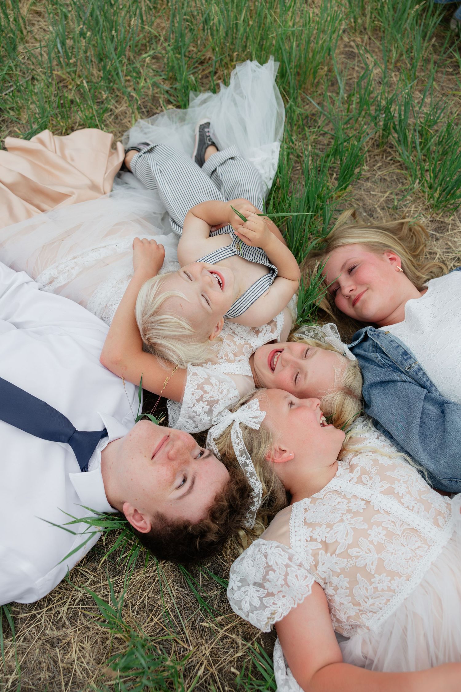 Five children in formal attire lying in a circle on grass, laughing and smiling together outdoors.