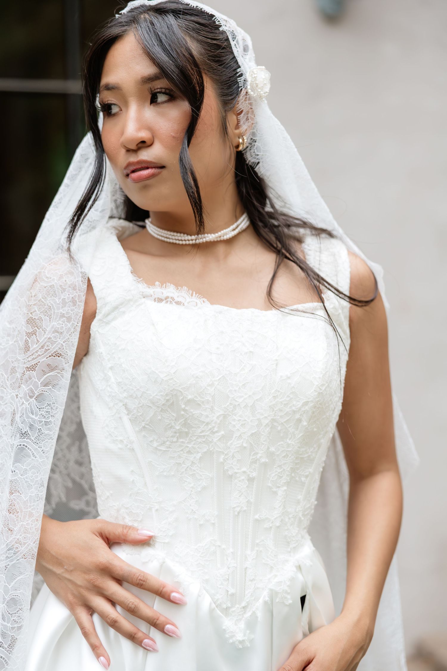Elegant Asian bride in white lace wedding dress and veil with pearl necklace posing outdoors.