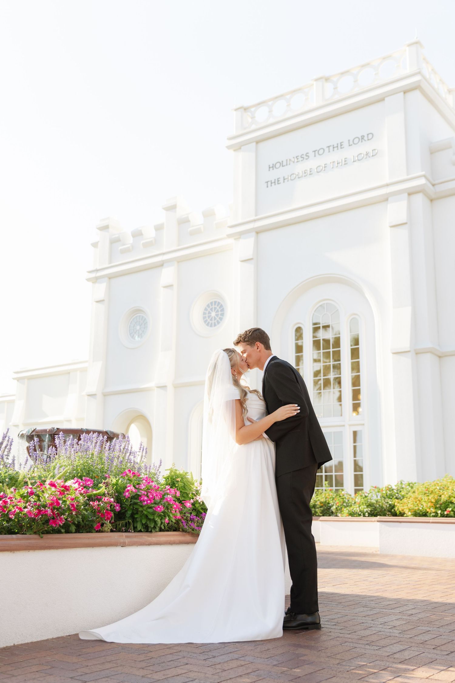 Bride and groom kissing in front of a white LDS temple surrounded by colorful flowers on their wedding day.