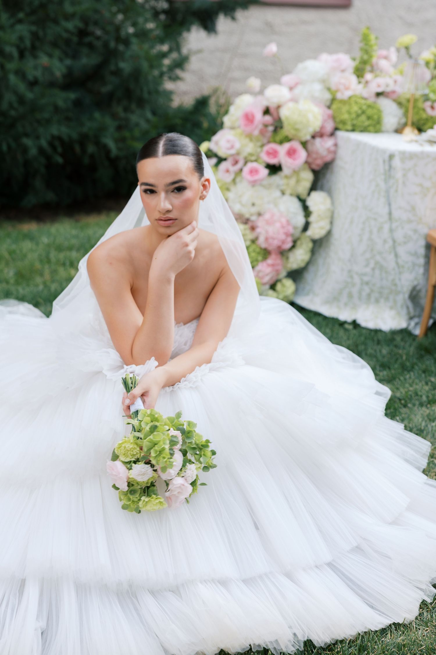 Elegant bride in a white tulle ballgown sits outdoors holding a bouquet, with floral arrangements in the background.