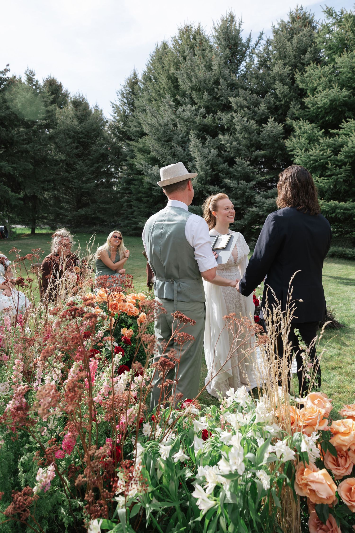 Bride and groom exchange vows outdoors surrounded by lush colorful garden flowers and tall pine trees.