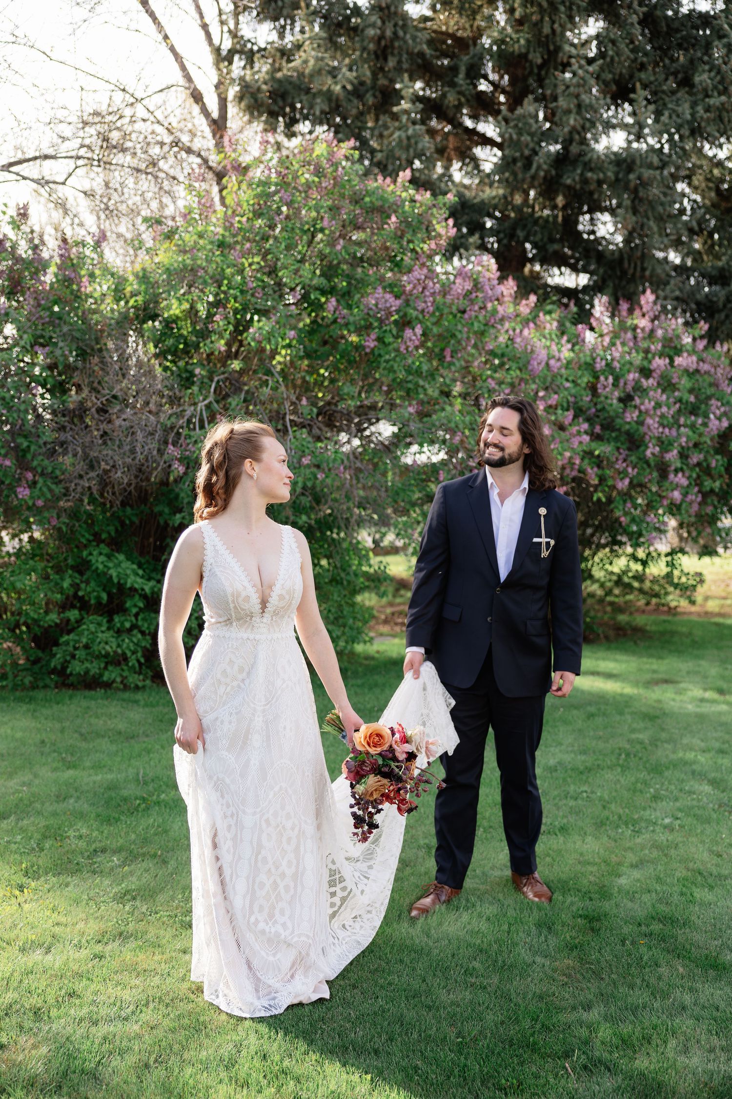 Bride and groom walking together in lush garden with blooming lilac trees in background.
