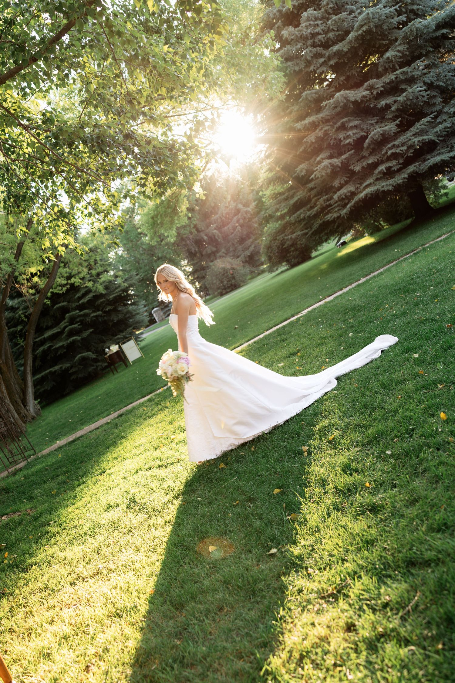 Bride in white gown with train walks through sunlit green park holding bouquet on her wedding day.