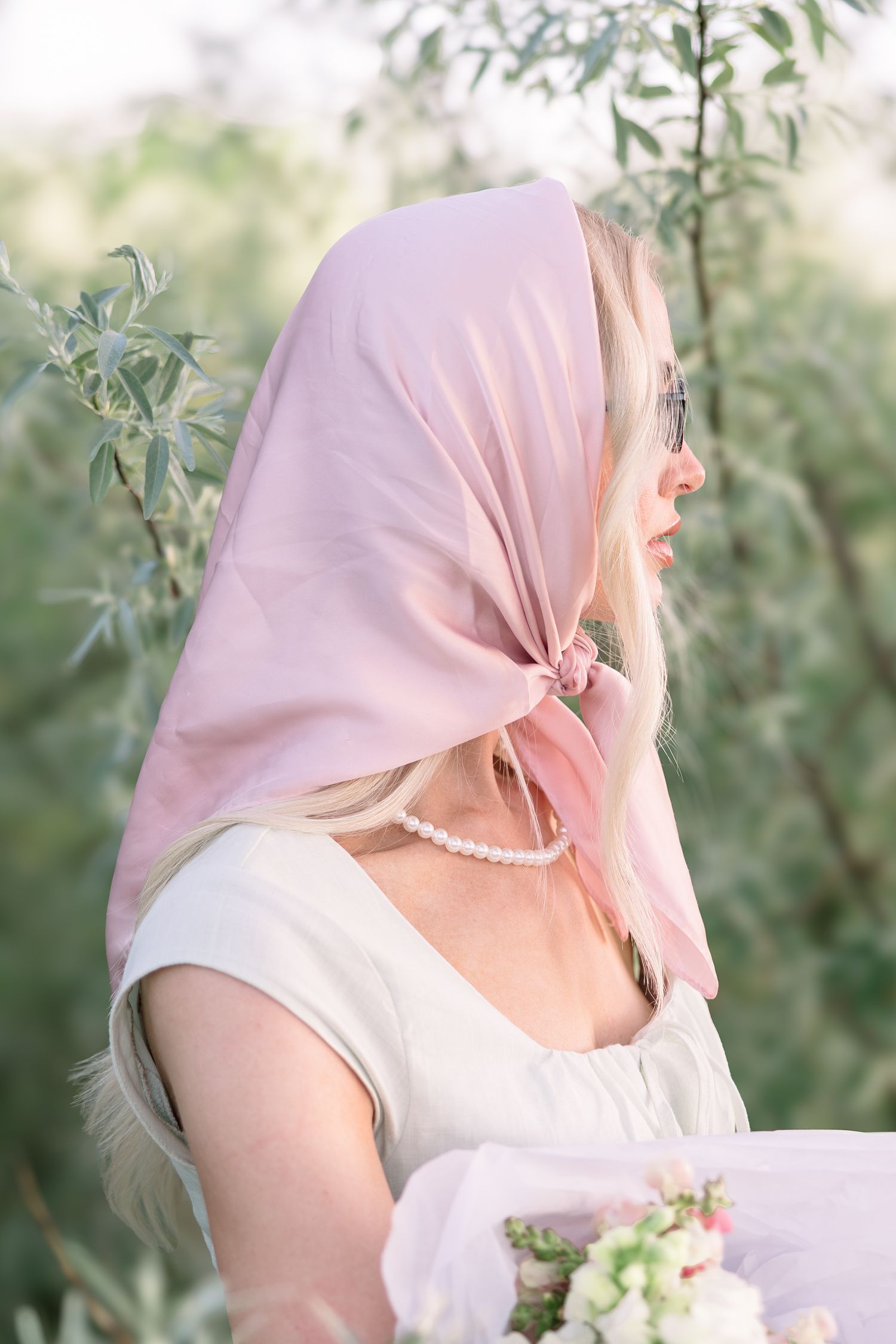 Blonde woman in pink head scarf, sunglasses, pearl necklace, and white dress holds flowers outdoors.