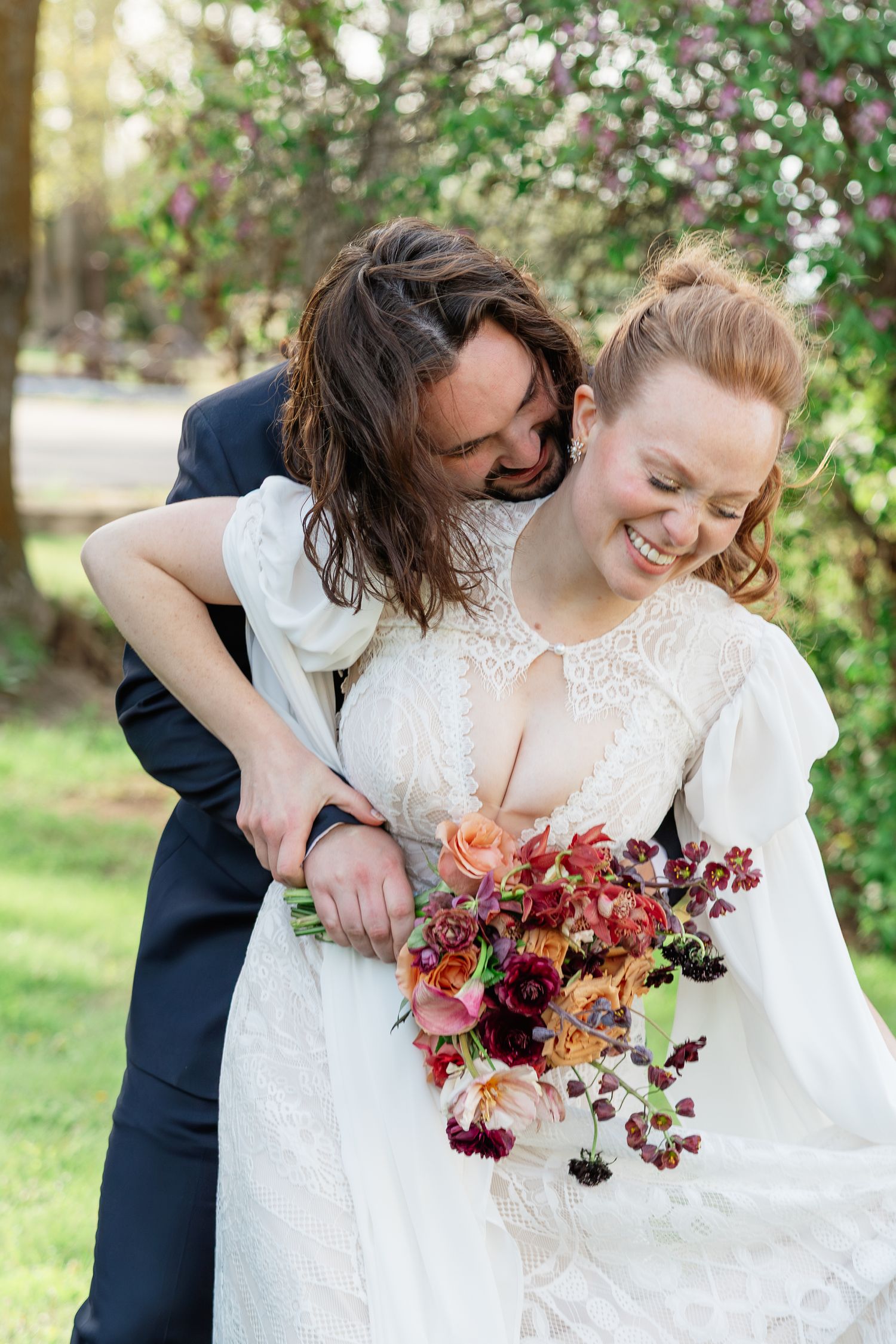 Joyful bride in lace gown laughs as groom embraces her from behind, holding a rich burgundy bridal bouquet.