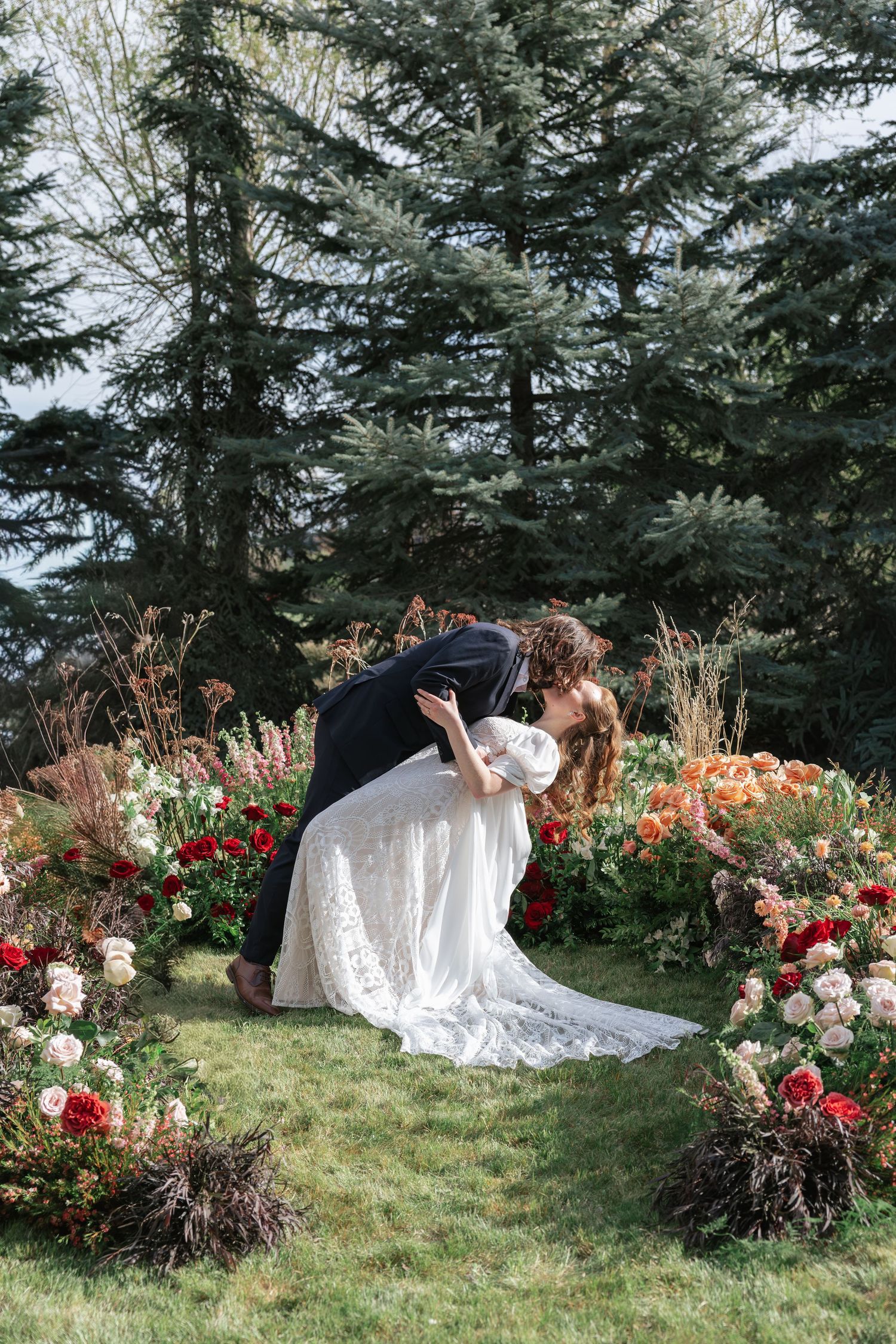 Bride and groom share a romantic dip kiss surrounded by a lush floral arch of red and pink blooms outdoors.