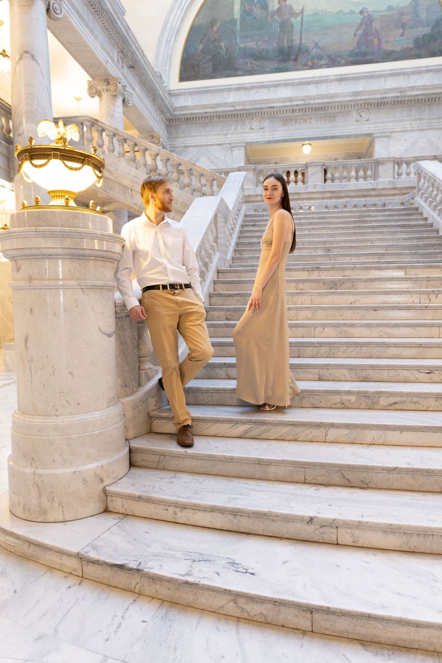 Elegant couple posing on grand marble staircase in ornate building, woman in beige gown, man in khaki outfit.