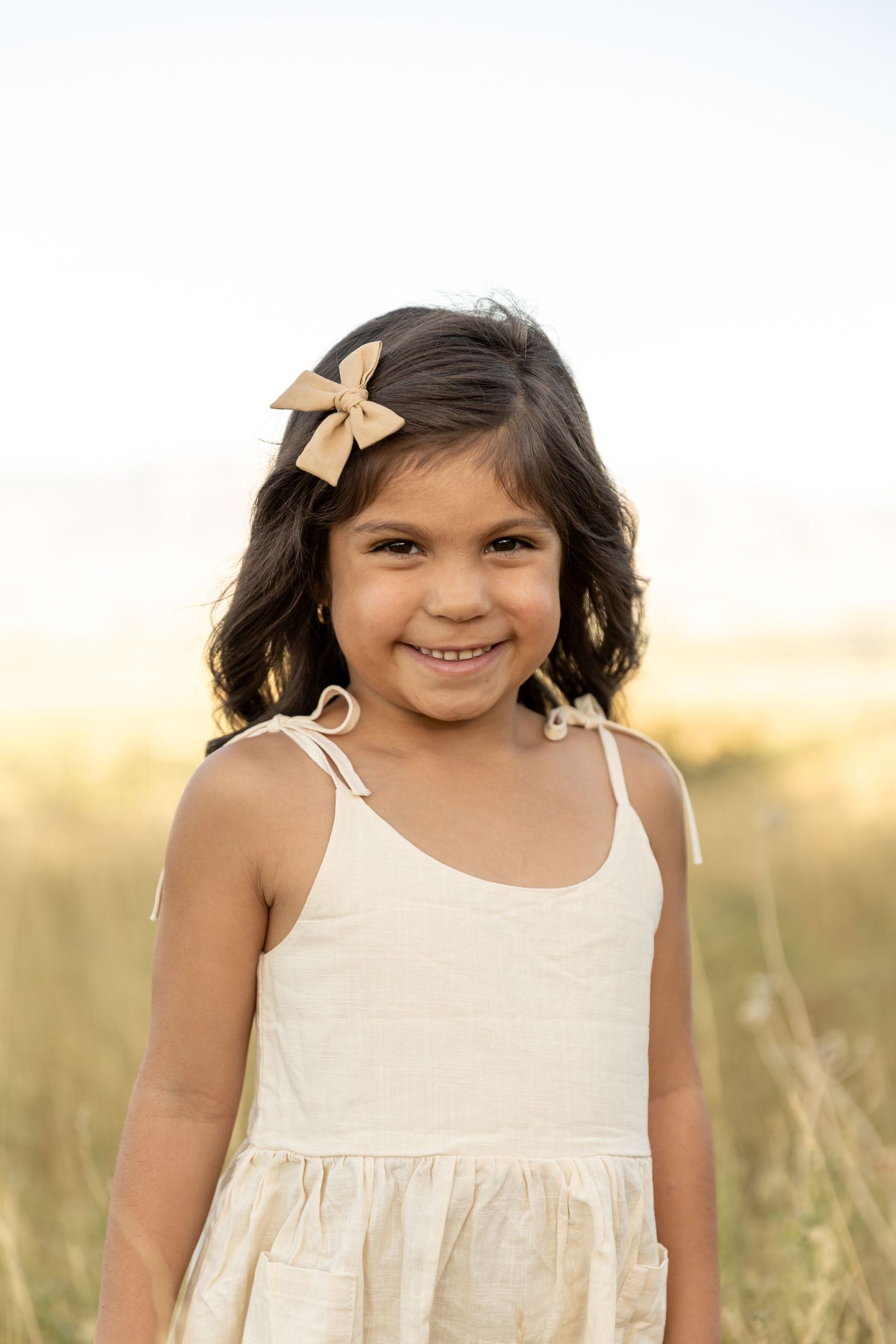 Smiling young girl with dark curly hair and a bow, wearing a cream sundress in a sunny field.