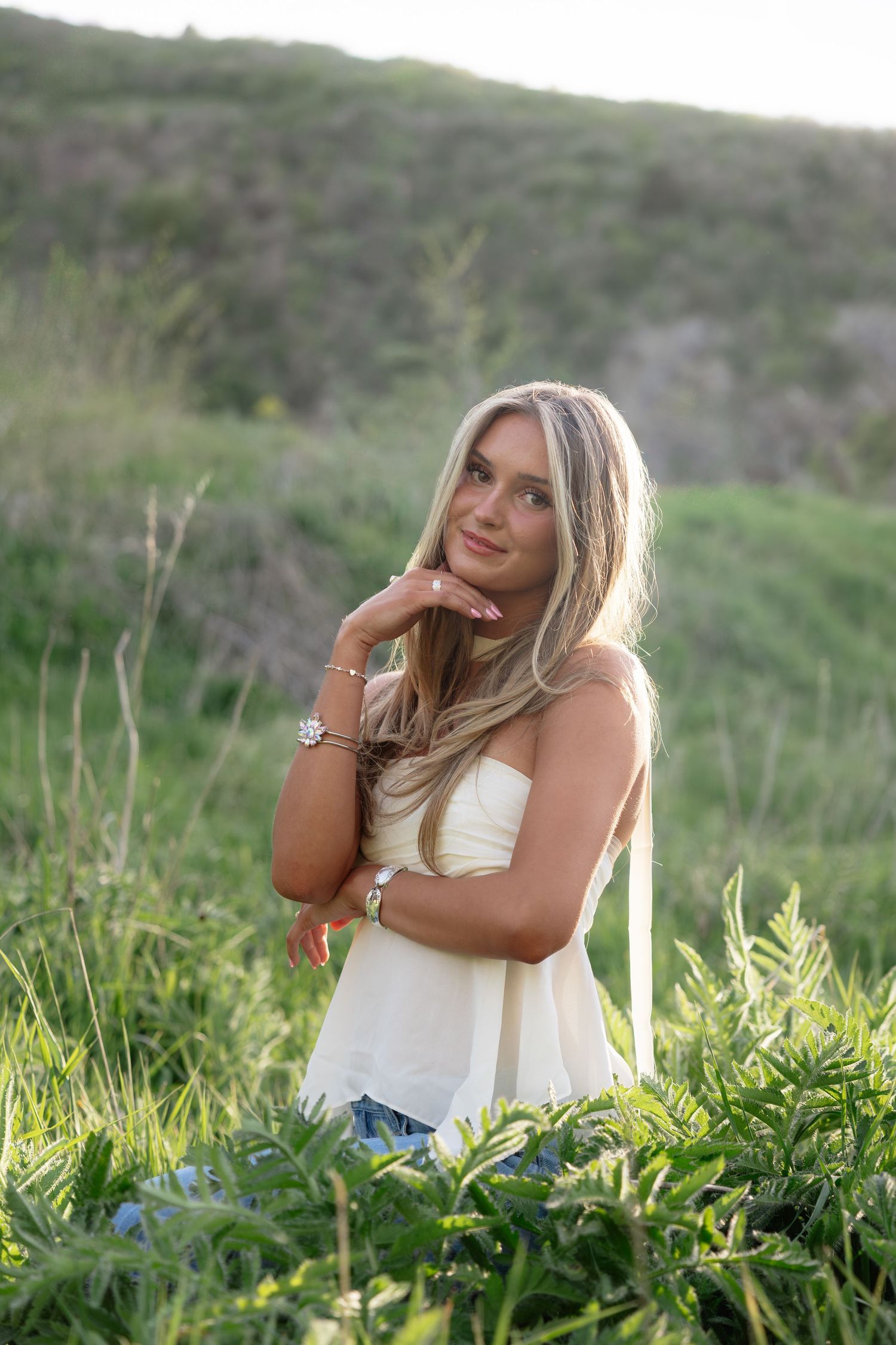 Young blonde woman in white top sitting in lush green field, smiling gracefully with hand on chin.