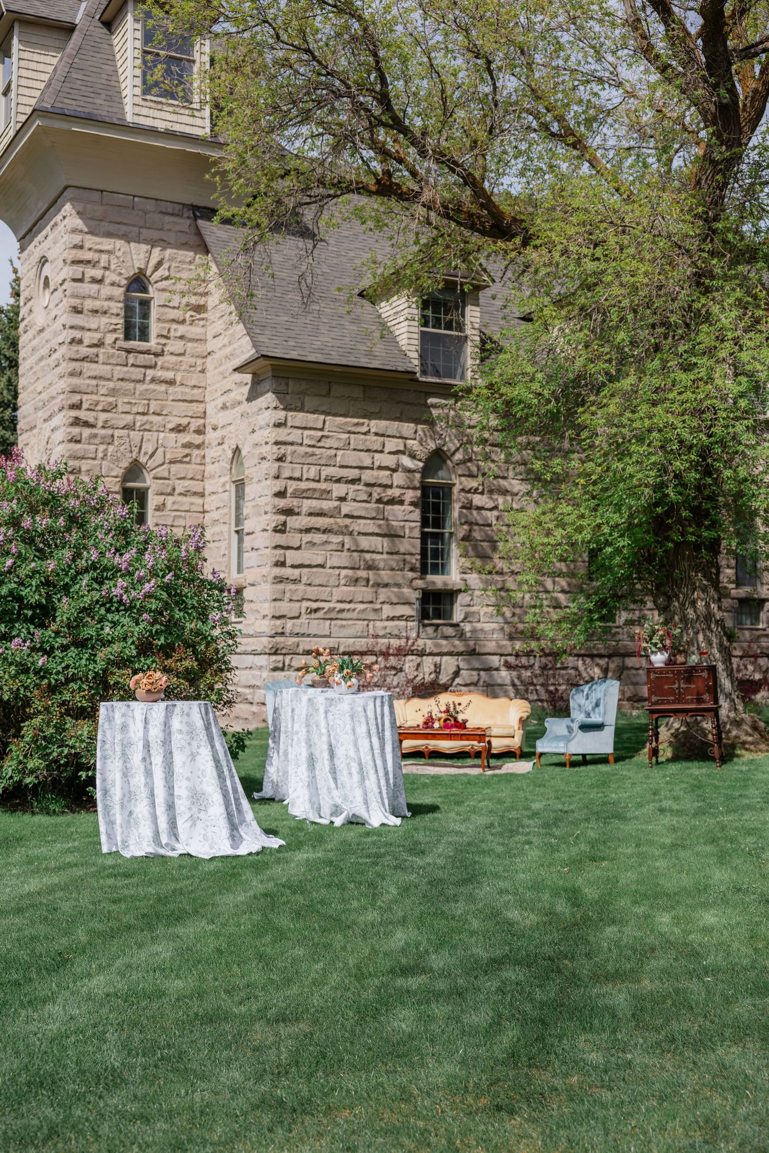 Elegant outdoor wedding reception setup with lace-covered cocktail tables and vintage furniture on a green lawn beside a stone building.