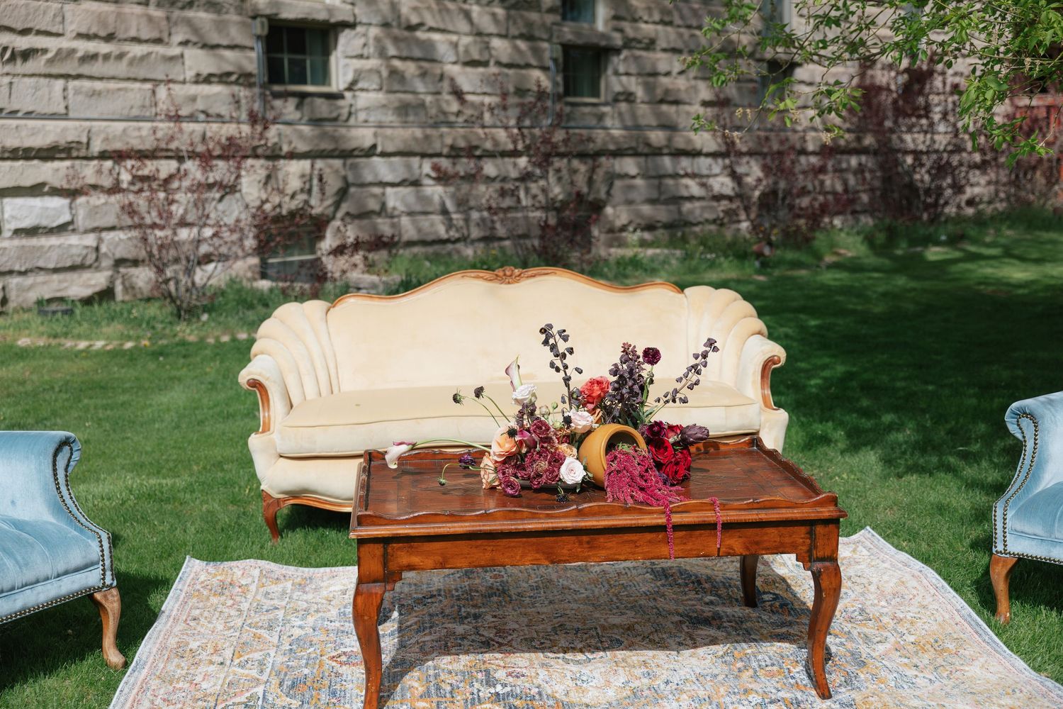 Vintage cream sofa with floral arrangement on wooden coffee table atop ornate rug at outdoor wedding lounge.