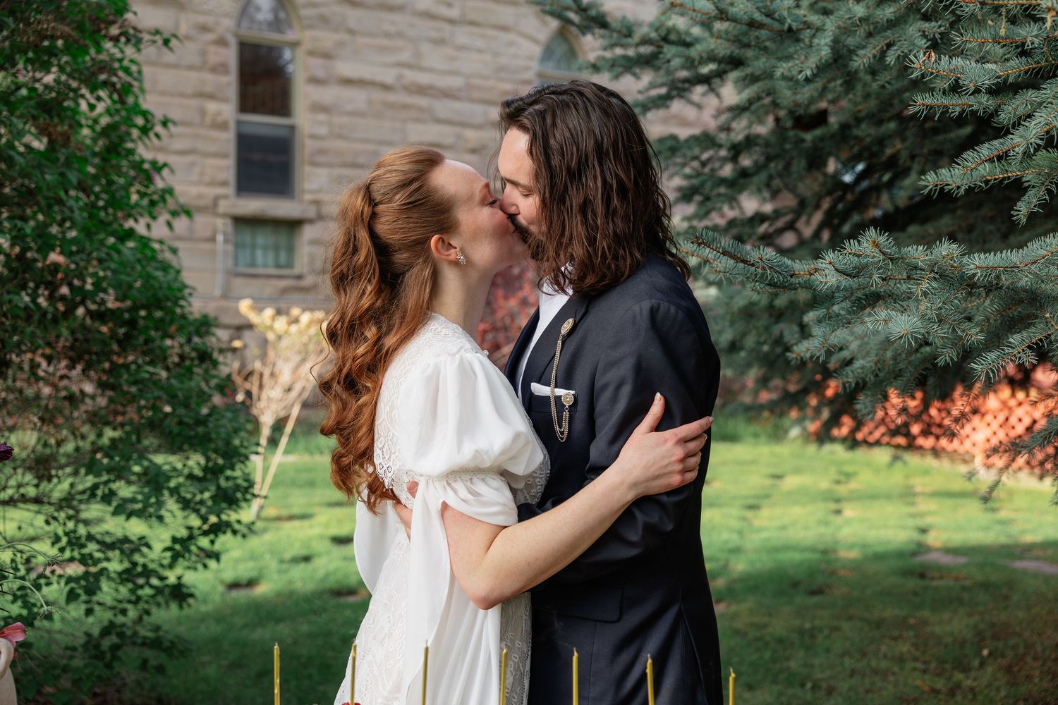 Newlyweds share first kiss embracing in lush garden with historic stone building in background.