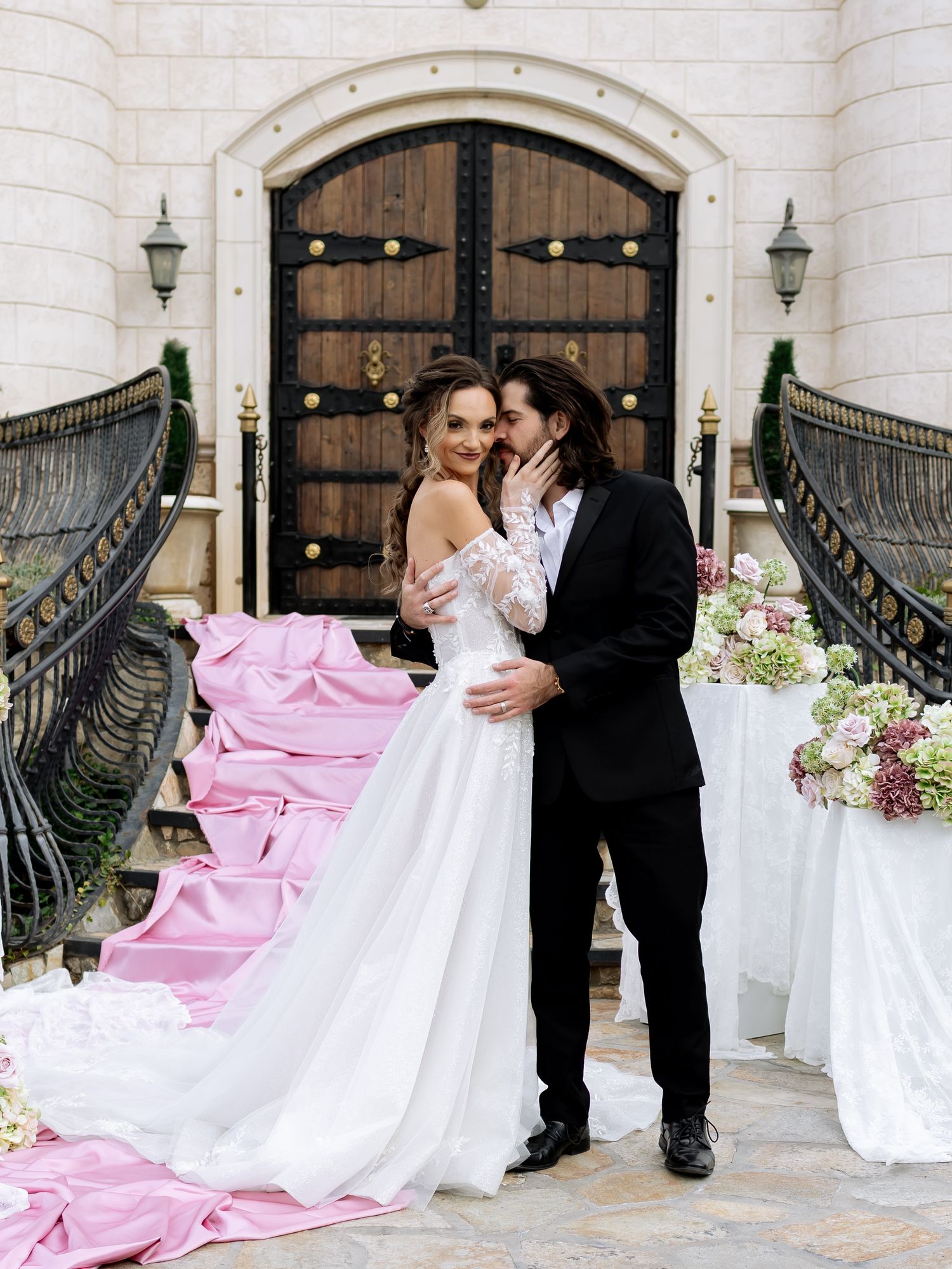 Bride in white gown with pink train embraces groom in black suit on elegant stone steps before ornate wooden doors.