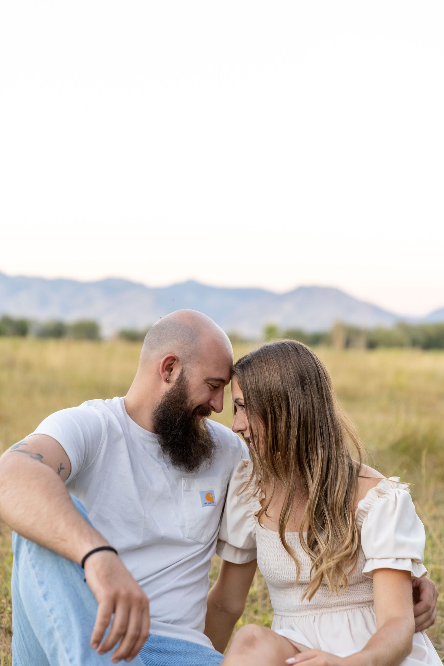 A couple sits close together in a golden field, touching foreheads romantically with mountains in the background.