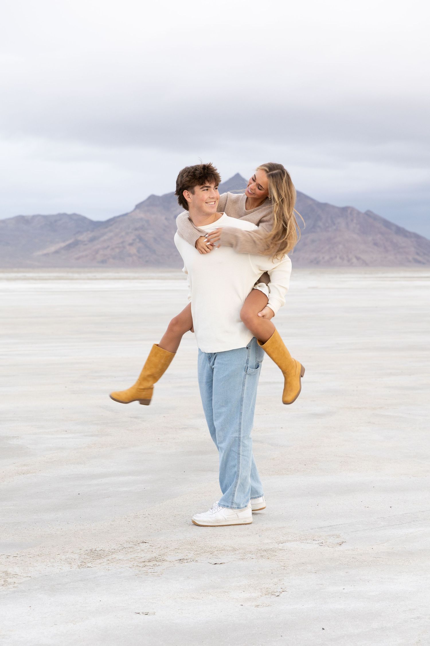 A young couple laughing as the man gives the woman a piggyback ride on the Bonneville Salt Flats with mountains behind.