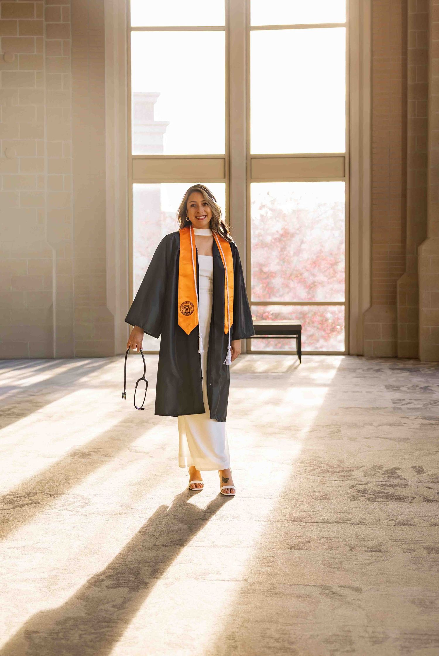 A smiling graduate walks in a sunlit hall wearing a black gown with an orange stole, holding a cap and stethoscope.