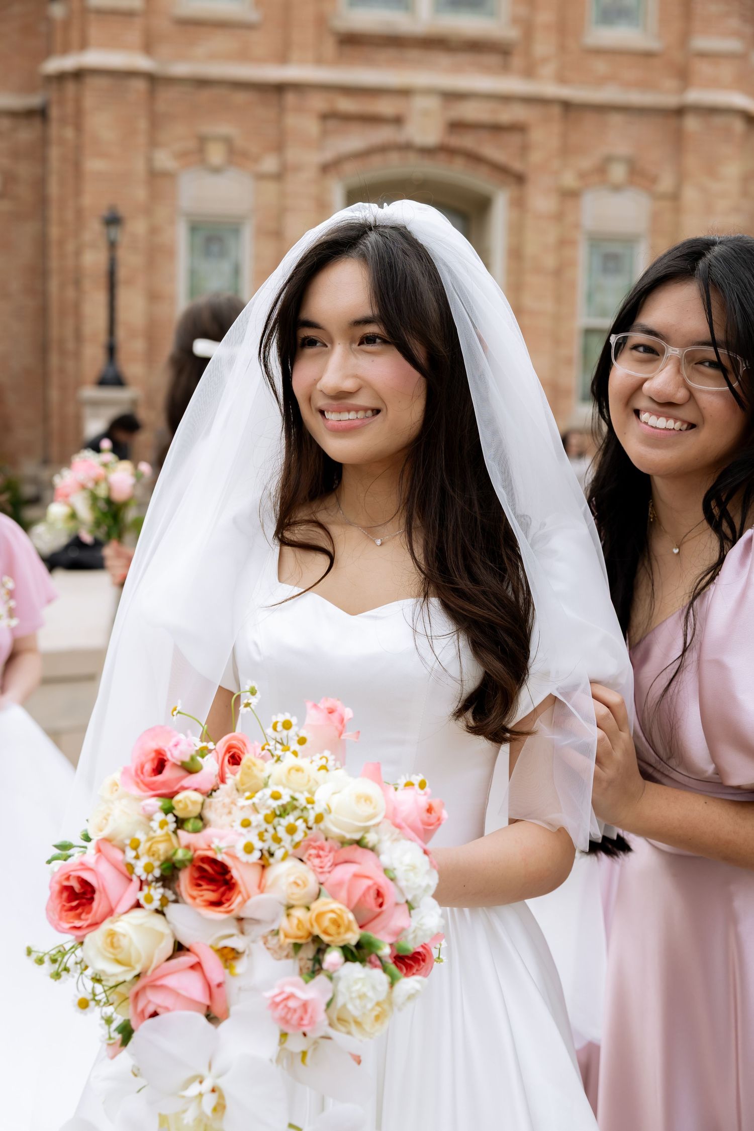 Smiling bride in white gown and veil holds pink bouquet beside bridesmaid in pink dress outdoors. Provo City Center Temple