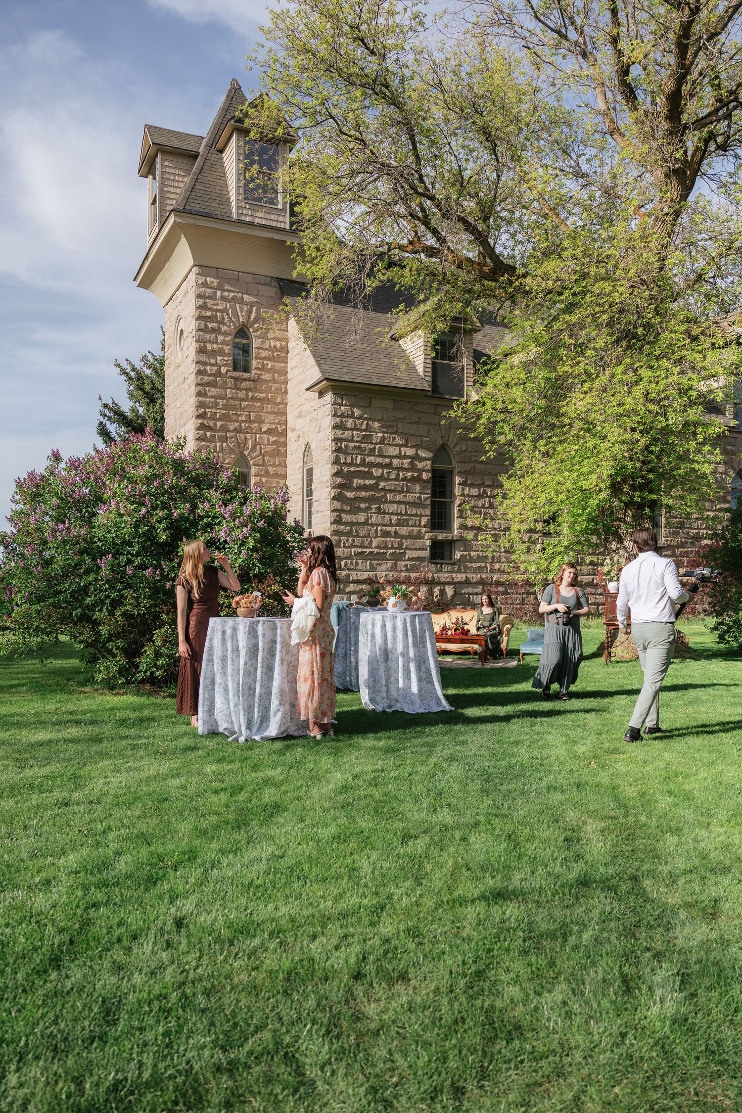 Wedding guests mingling outdoors near stone chapel with cocktail tables on green lawn.