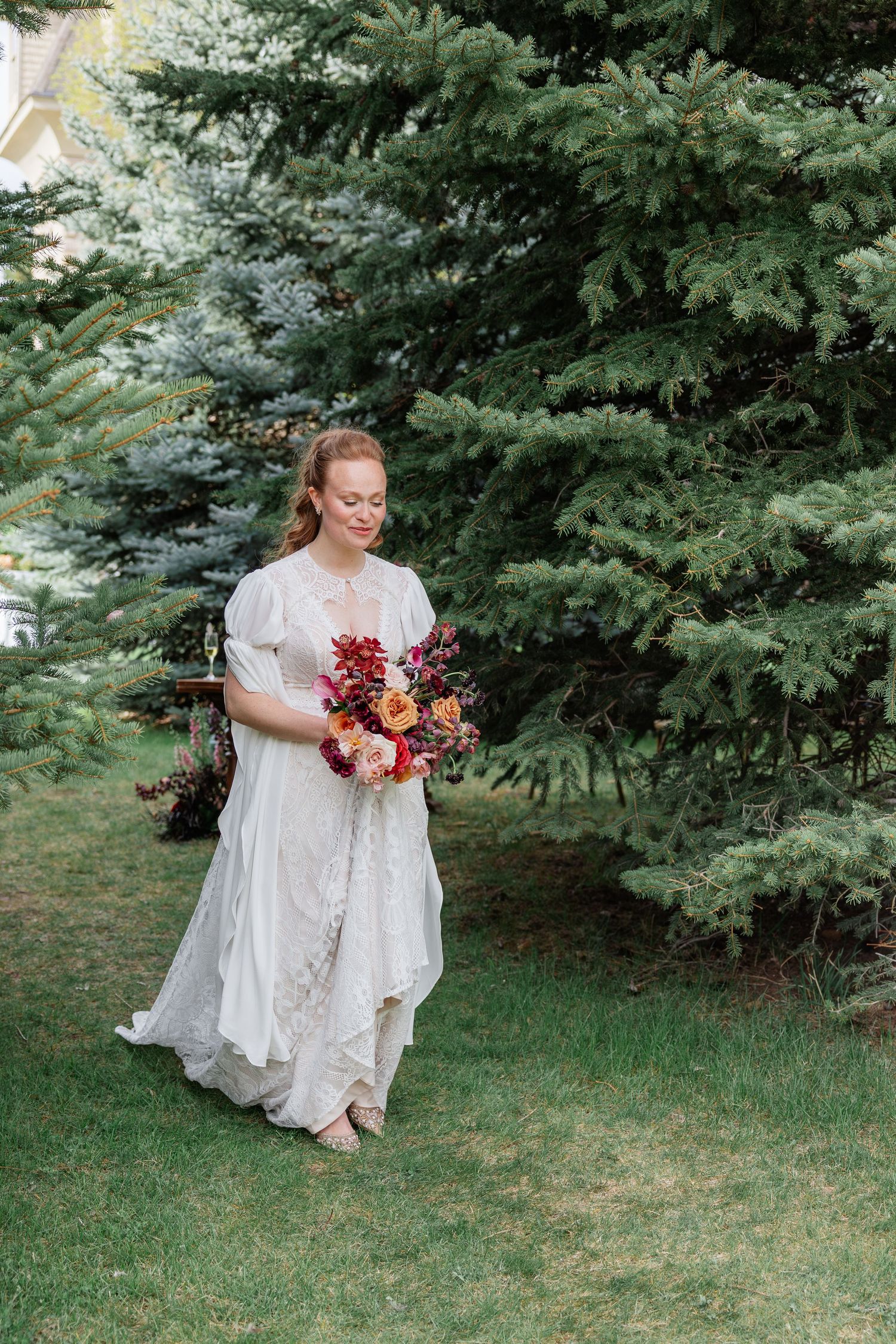 Bride in white lace dress walks through garden holding vibrant red and orange bridal bouquet.