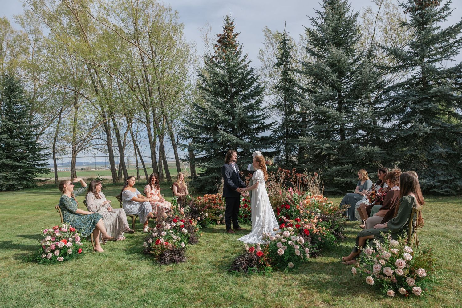 Intimate outdoor wedding ceremony on lush green lawn with floral arrangements and tall evergreen trees in background.