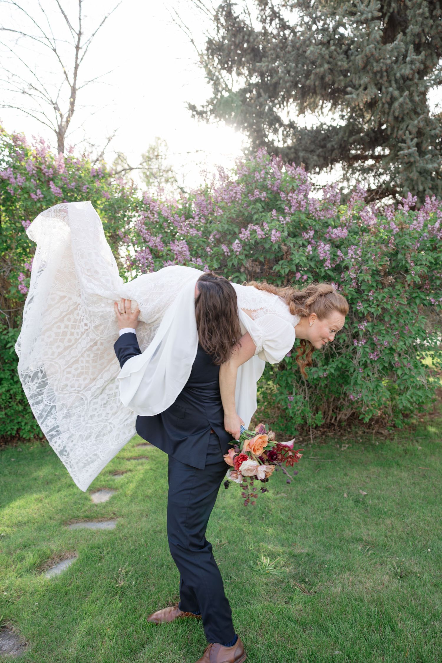 Groom playfully carries bride over his shoulder in a lush garden with blooming lilacs and bouquet.