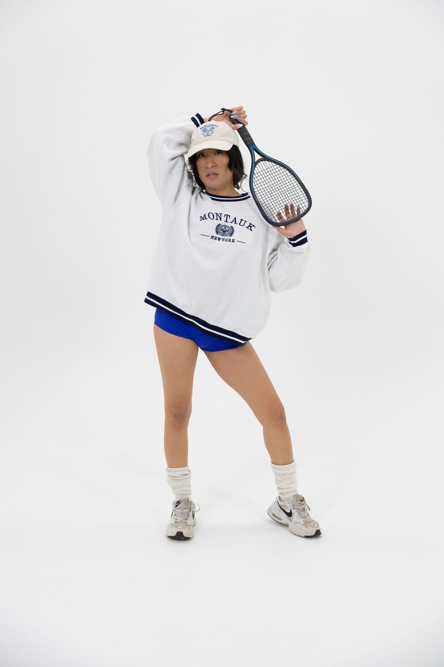 Woman posing with tennis racket wearing white Montauk sweatshirt, blue shorts, bucket hat, and Nike sneakers.