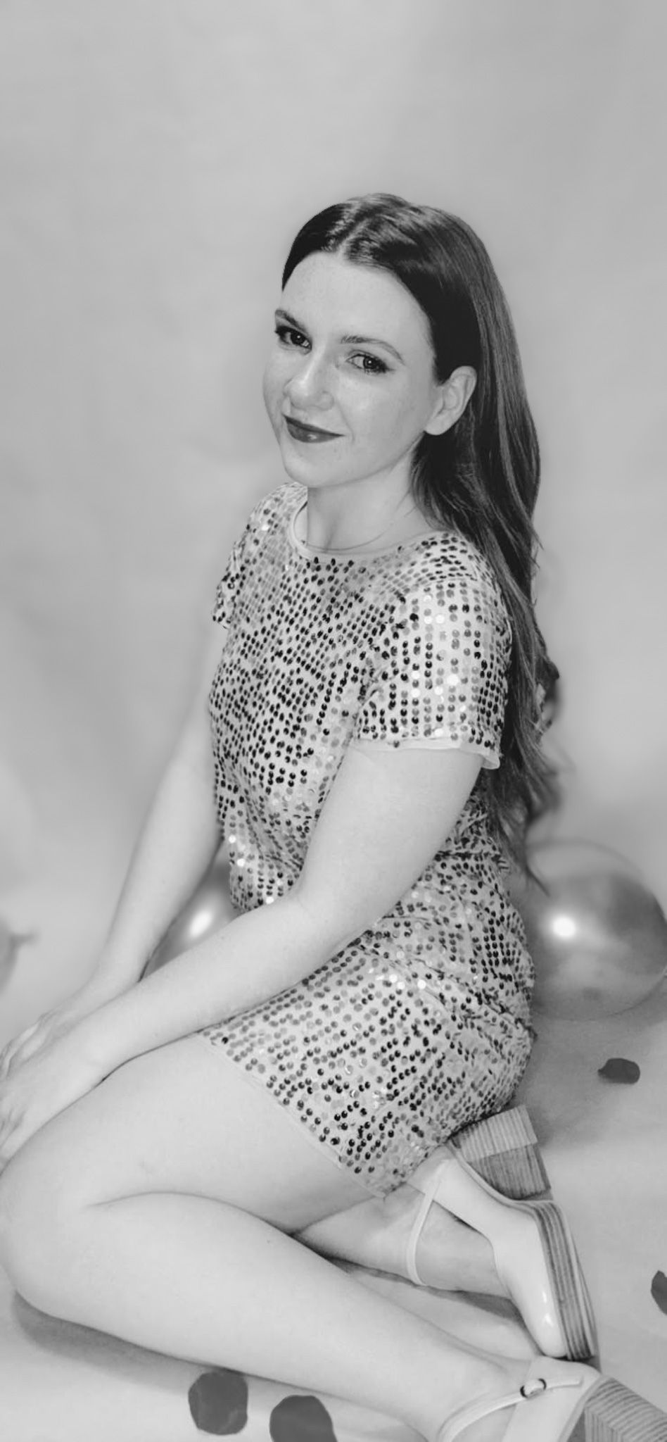 Black and white photo of a young woman in a sequined mini dress sitting gracefully with balloons in the background.
