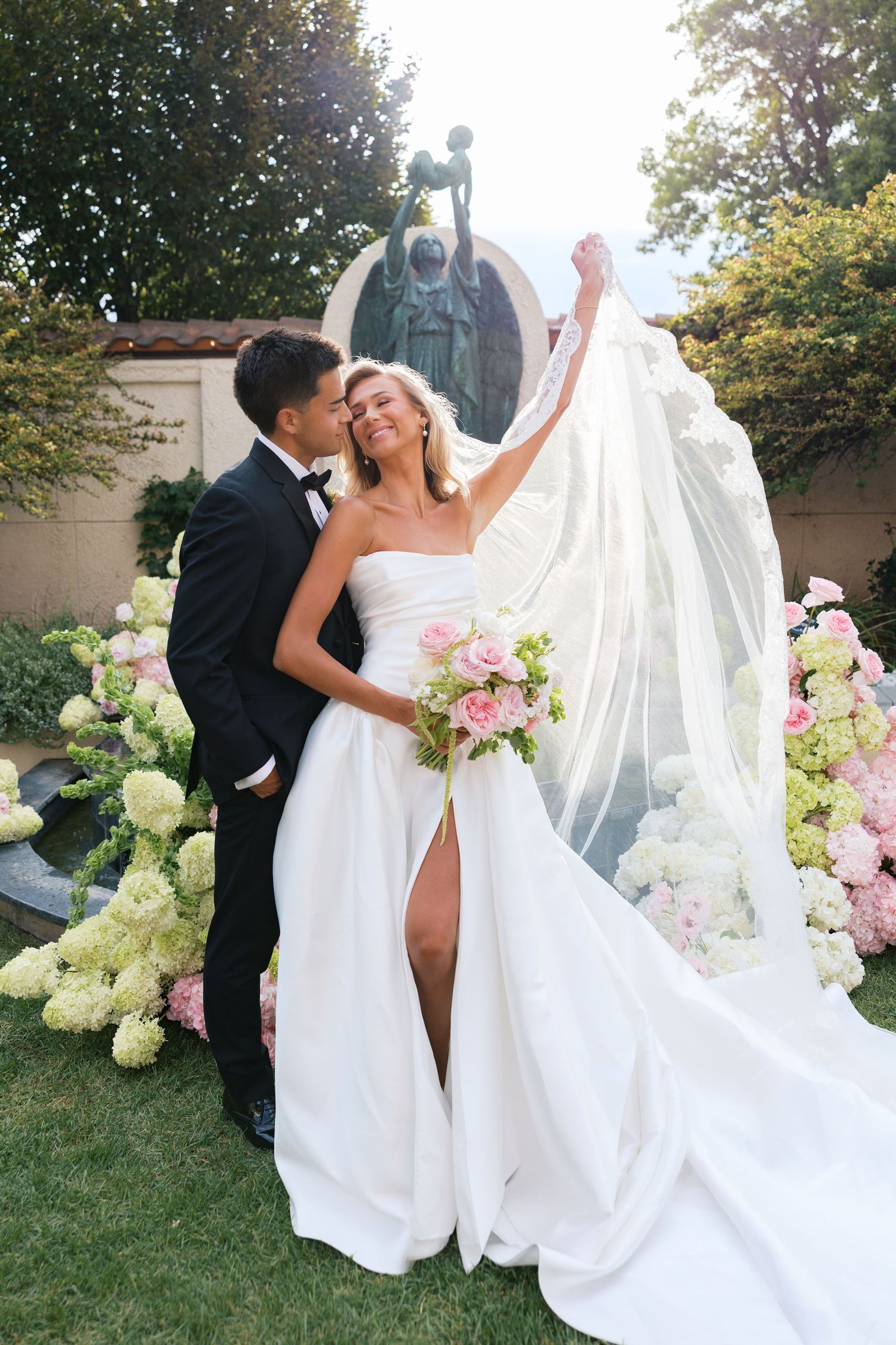 Bride in strapless white gown with flowing veil holds bouquet as groom in tuxedo kisses her cheek outdoors.