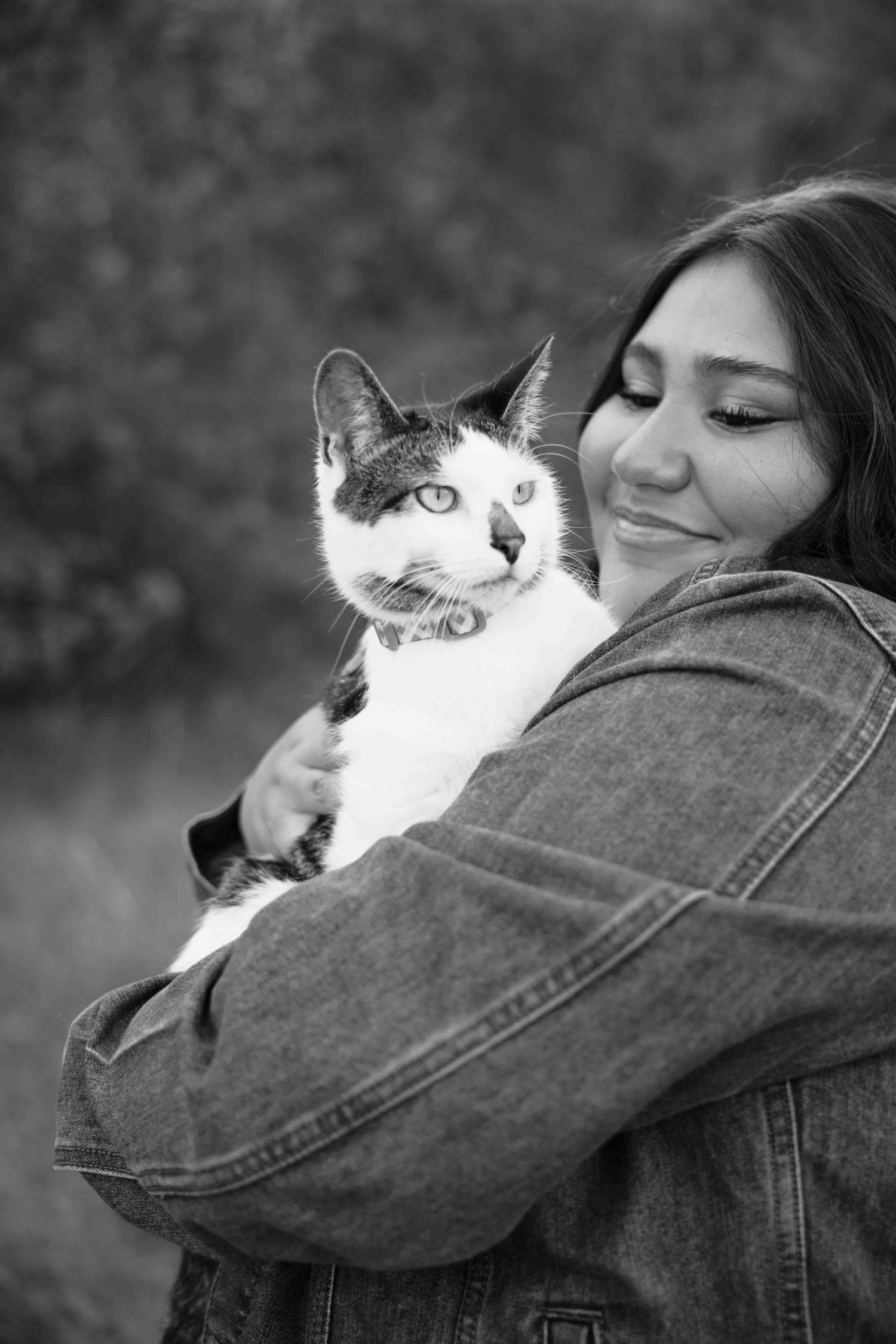 Black and white photo of a smiling young woman in denim jacket tenderly holding a tabby cat outdoors.