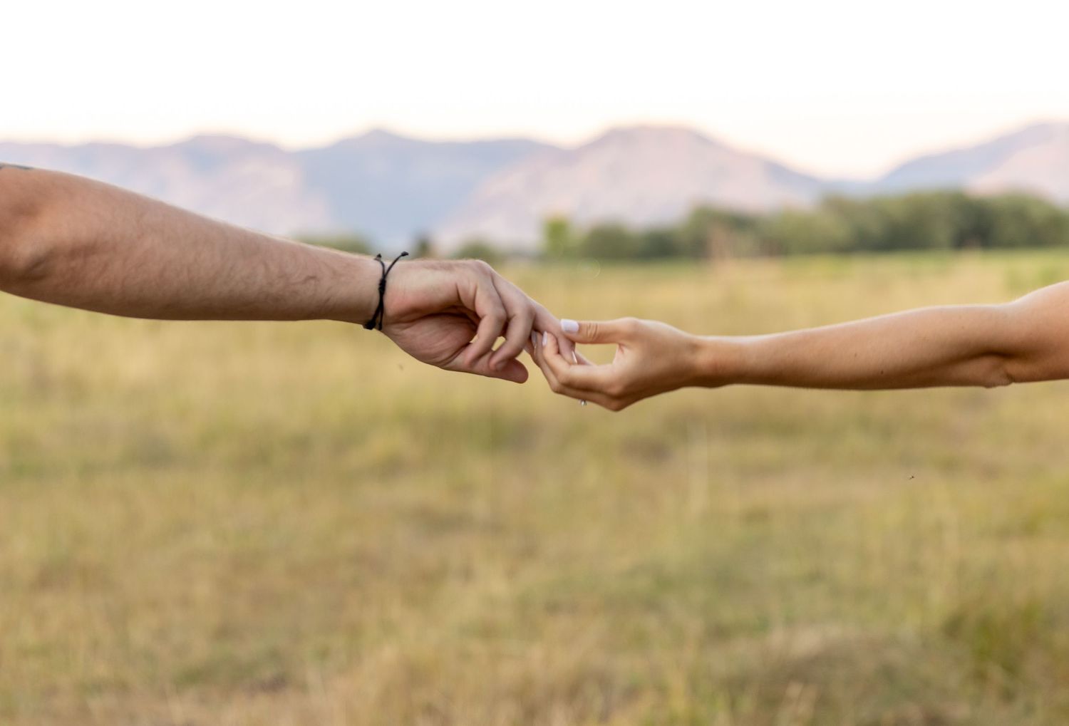 Two people gently touching fingertips in a field with mountains in the background, symbolizing connection and love.