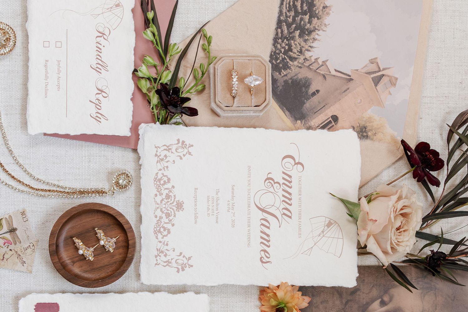 Elegant wedding stationery flatlay with floral invitations, ring box, earrings, and greenery accents on white background.