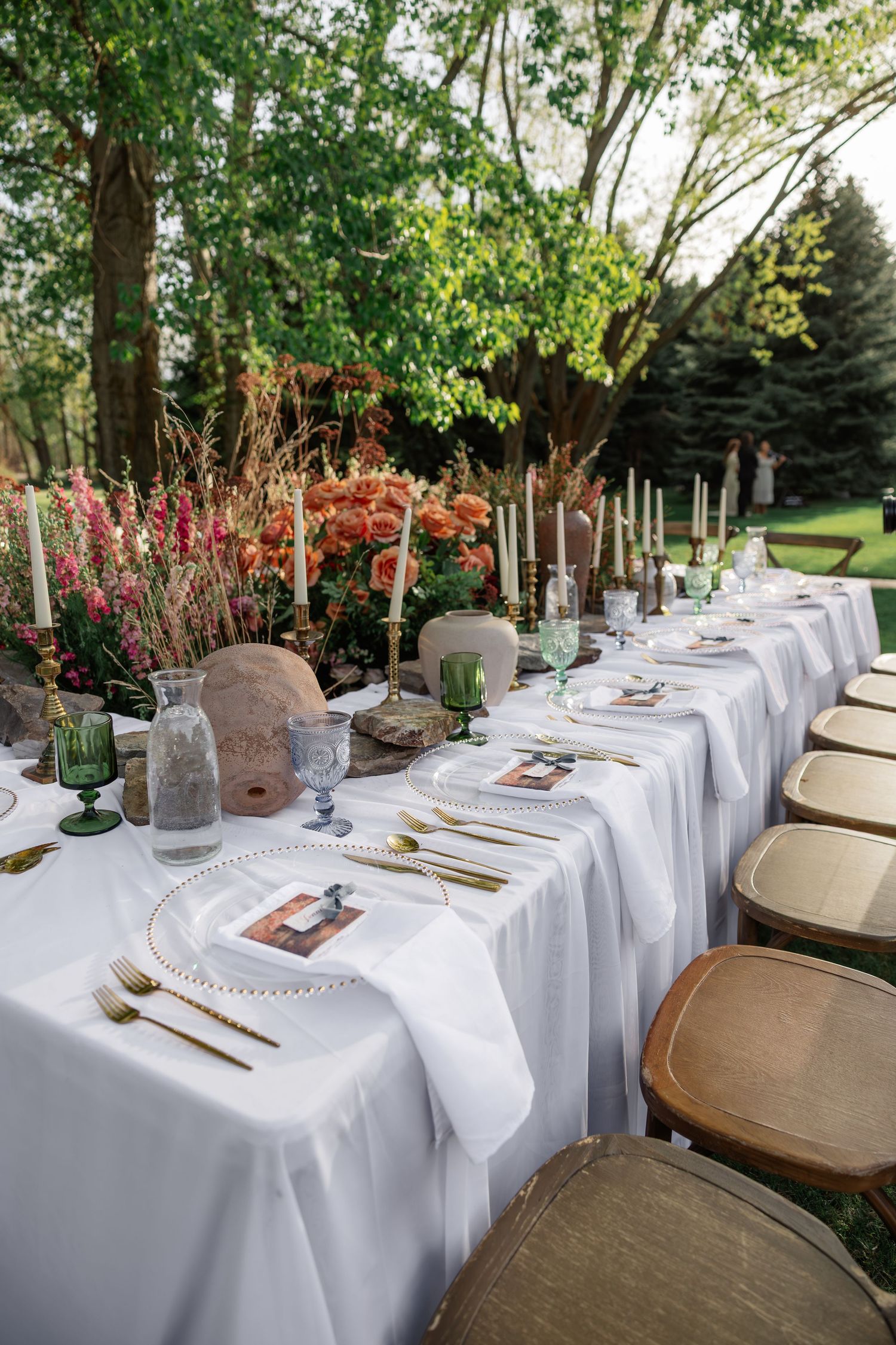 Elegant outdoor wedding reception table with white linen, gold cutlery, candles, and vibrant floral centerpieces.