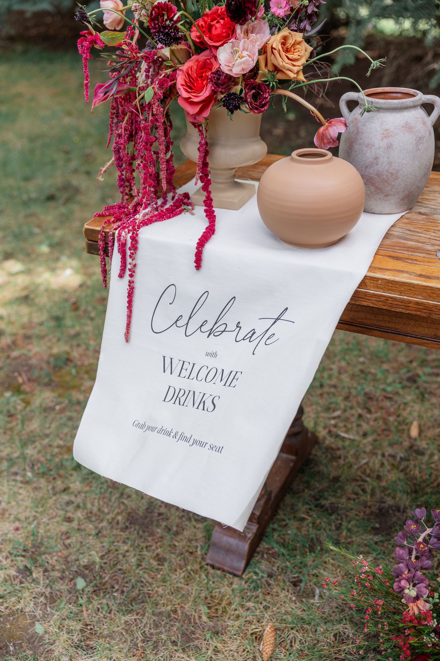 Rustic wooden table with celebrate welcome drinks sign, terracotta vases, and vibrant floral arrangement outdoors.