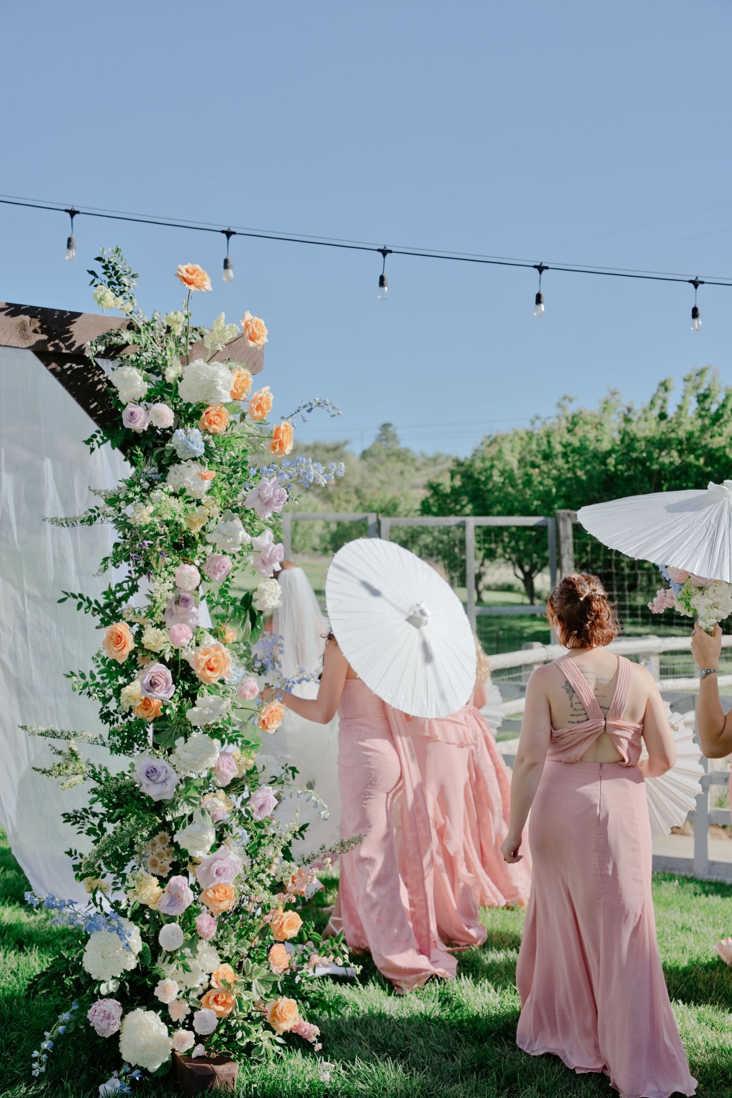 Two bridesmaids in pink dresses with parasols walk past a floral arch with peach and white roses at an outdoor wedding.