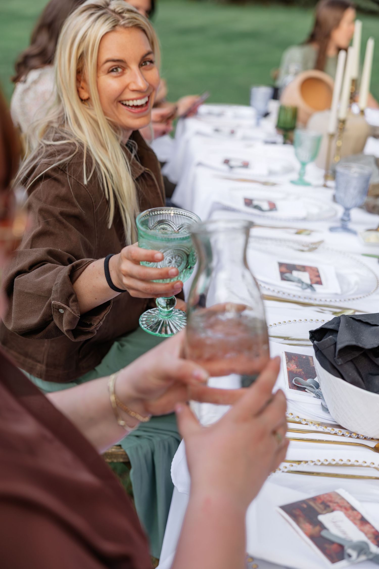 Smiling blonde wedding guest toasting with green glass at outdoor reception dinner table.
