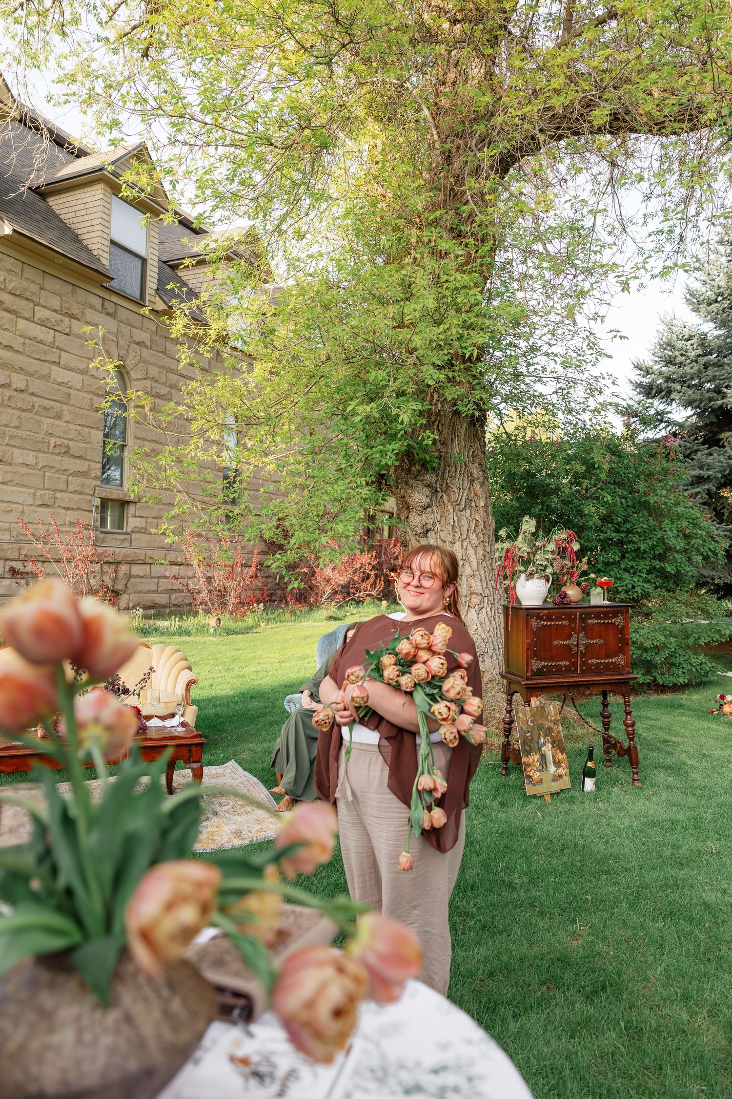 Woman holding large bundle of peach roses standing in lush garden near stone house.