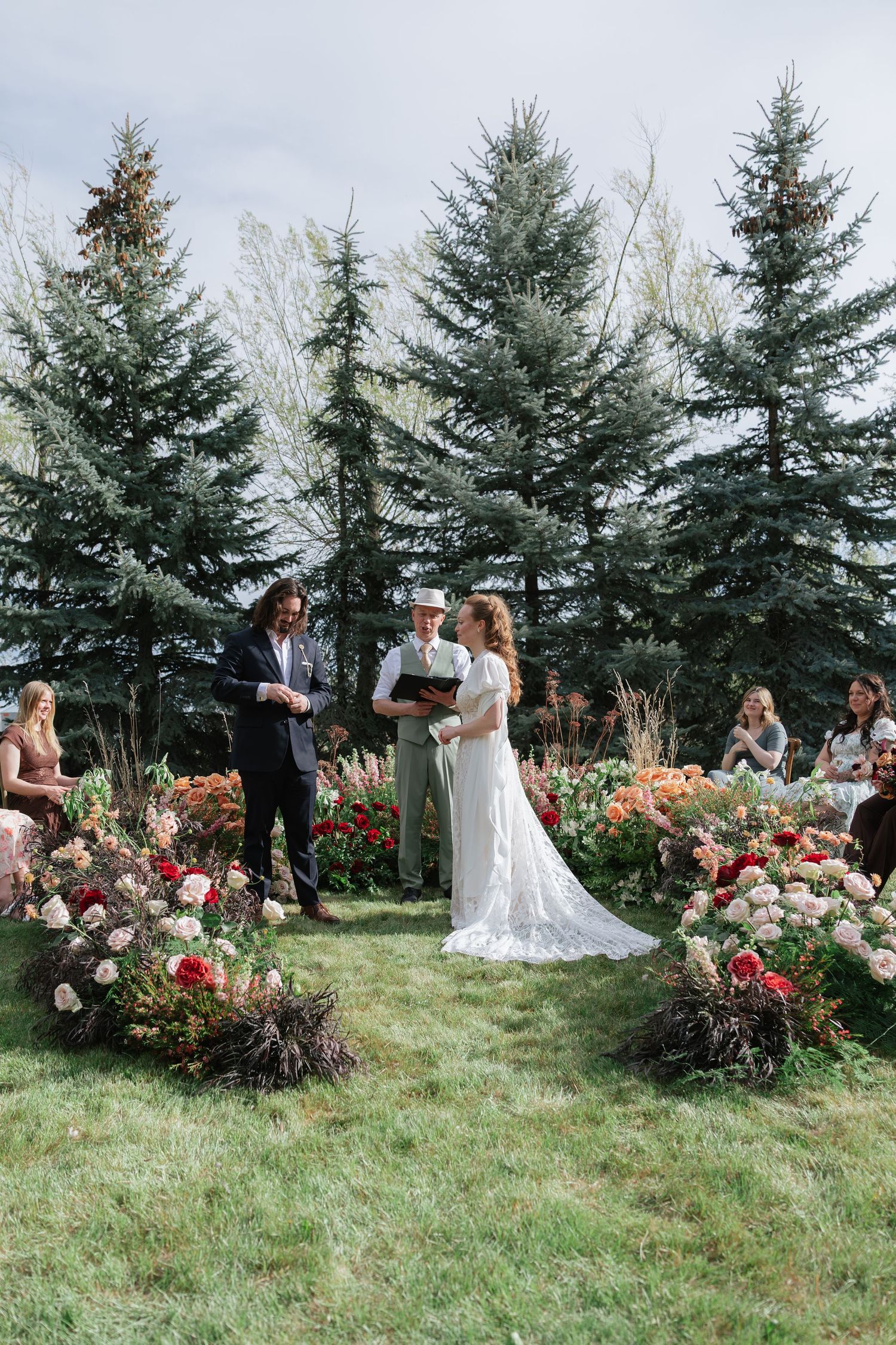 Outdoor wedding ceremony with couple exchanging vows amid lush floral arrangements and evergreen trees.