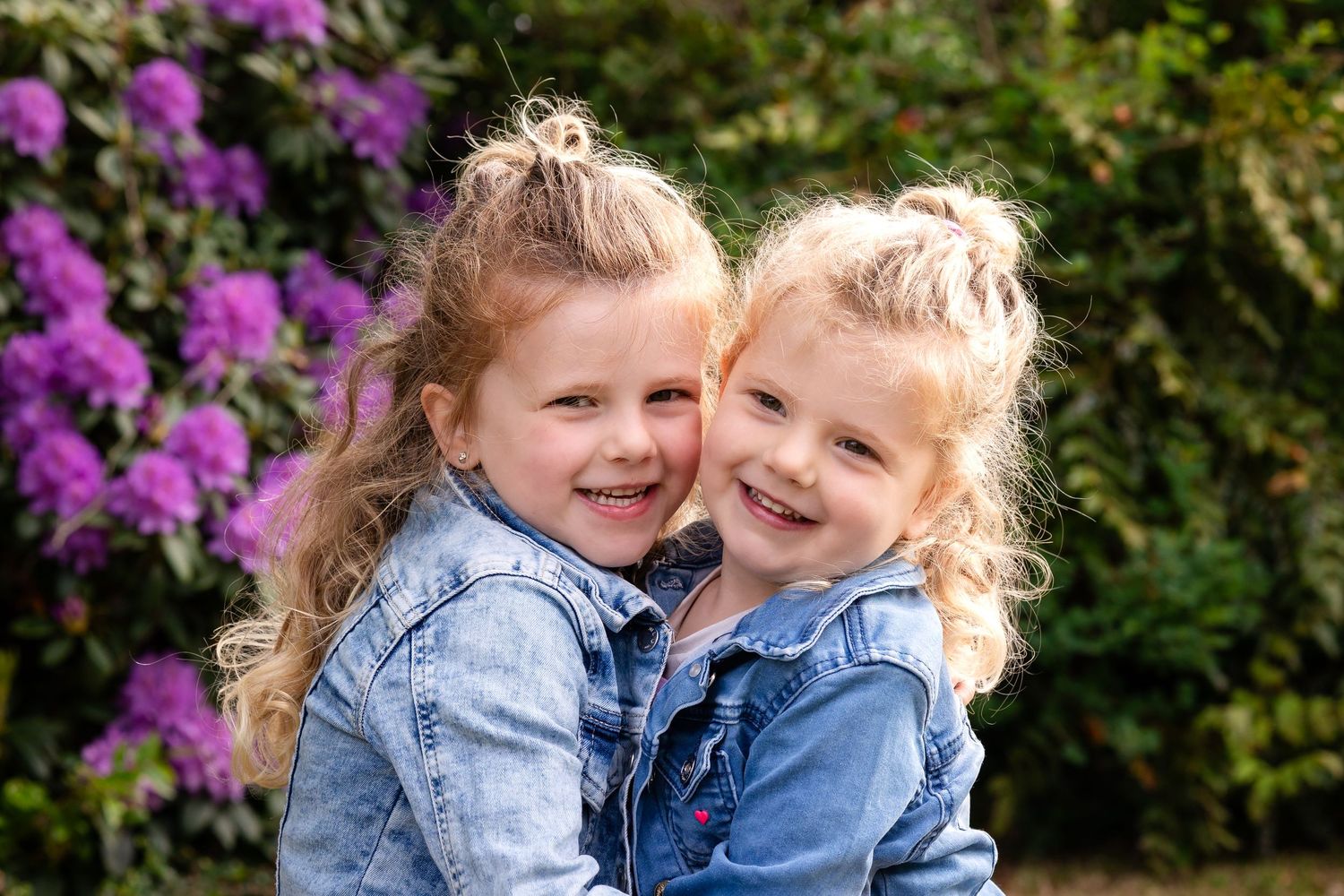 Two children wearing denim jackets share a sweet moment together in front of vibrant purple flowers.