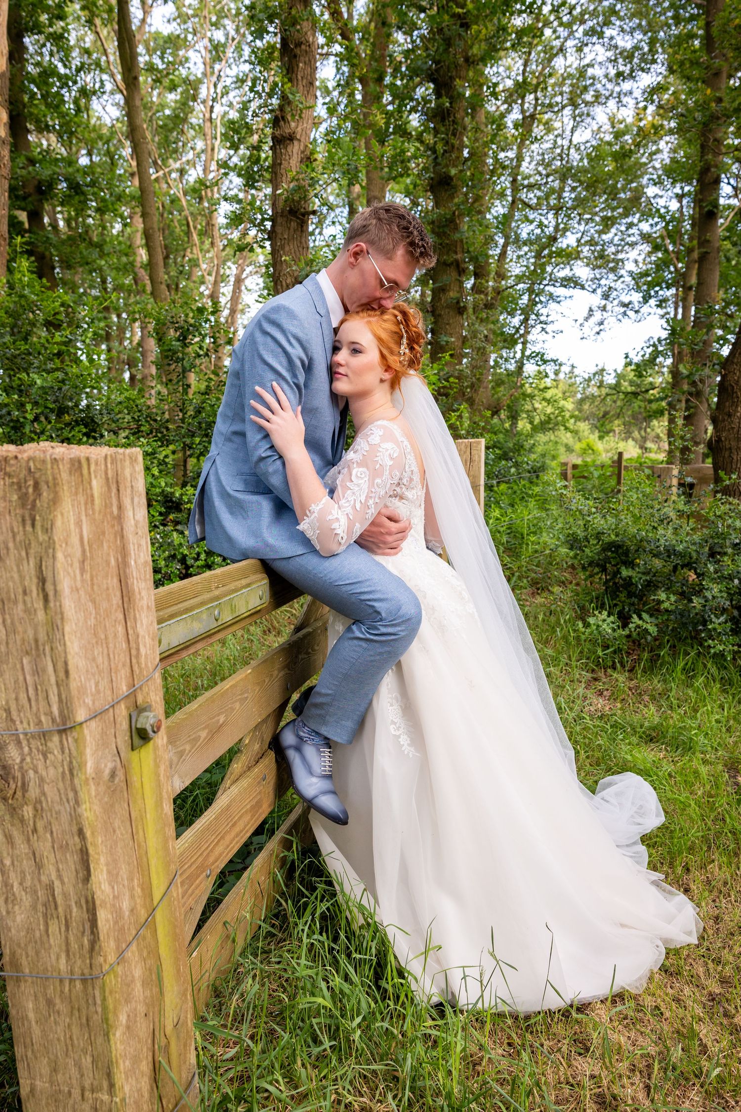 Romantic wedding photo shoot by a rustic wooden fence in a wooded area.
