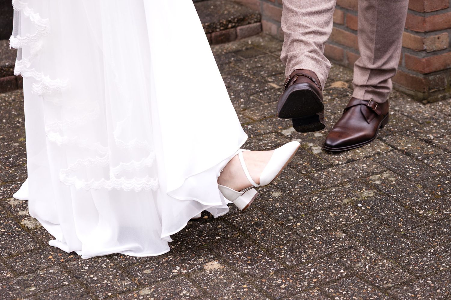 The hem of a white wedding dress and formal shoes visible against dark pavement.