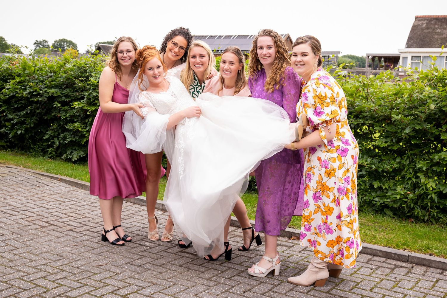 Bridesmaids in colorful dresses help adjust wedding gown on outdoor brick pathway.