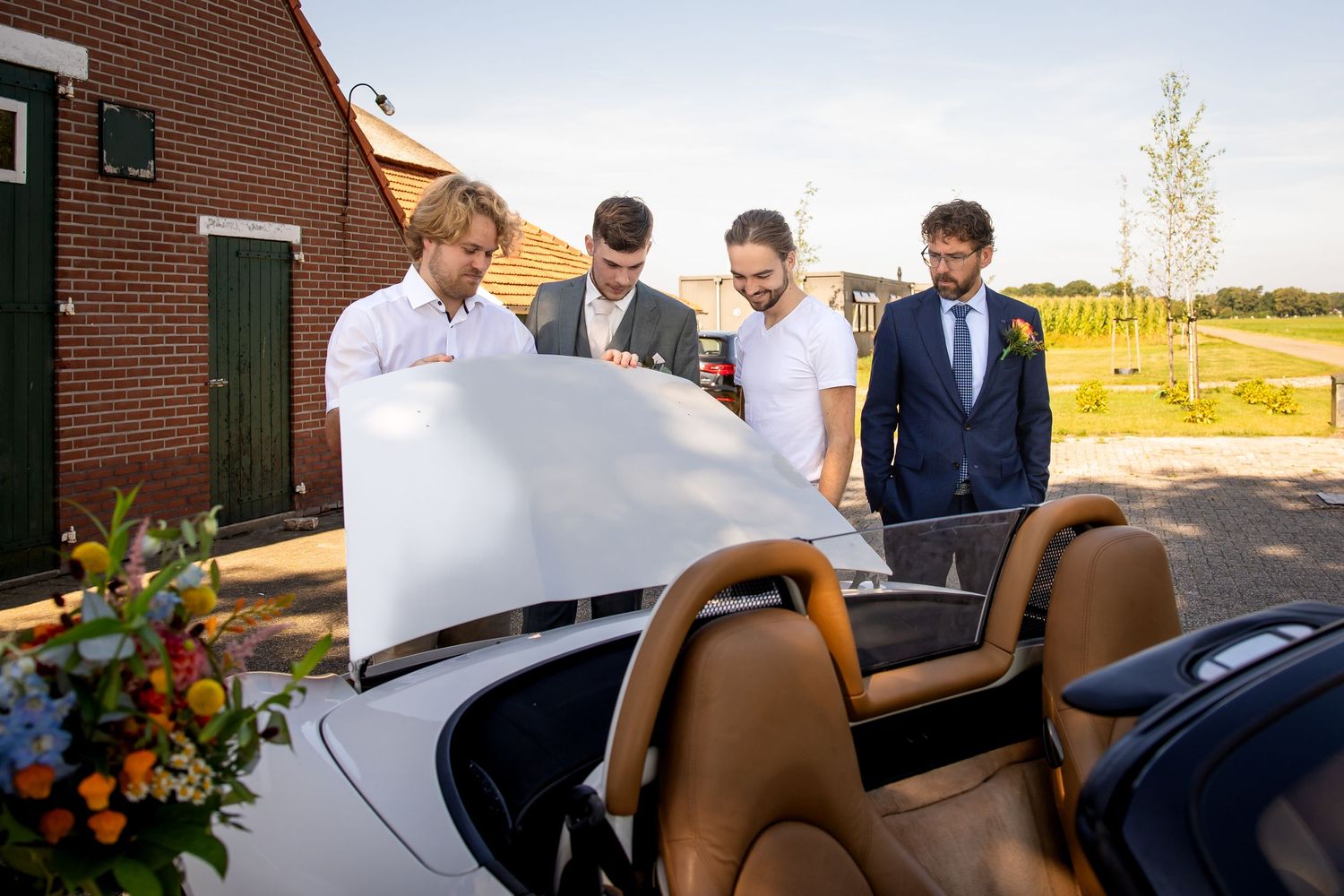 A group gathers around a convertible car outside a brick building.