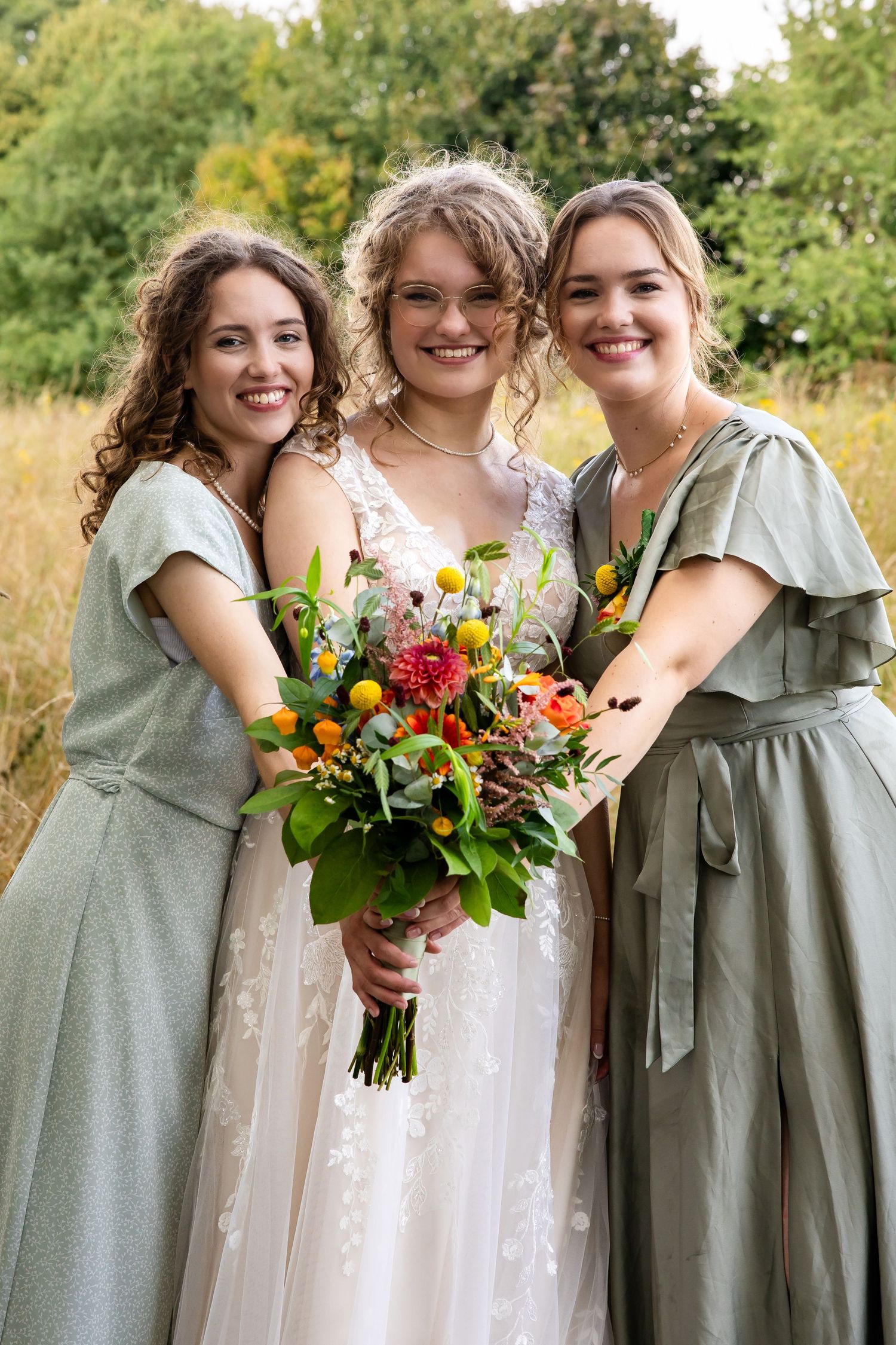 Bridesmaids in sage green dresses pose together with vibrant wildflower bouquets against a sunny outdoor backdrop.