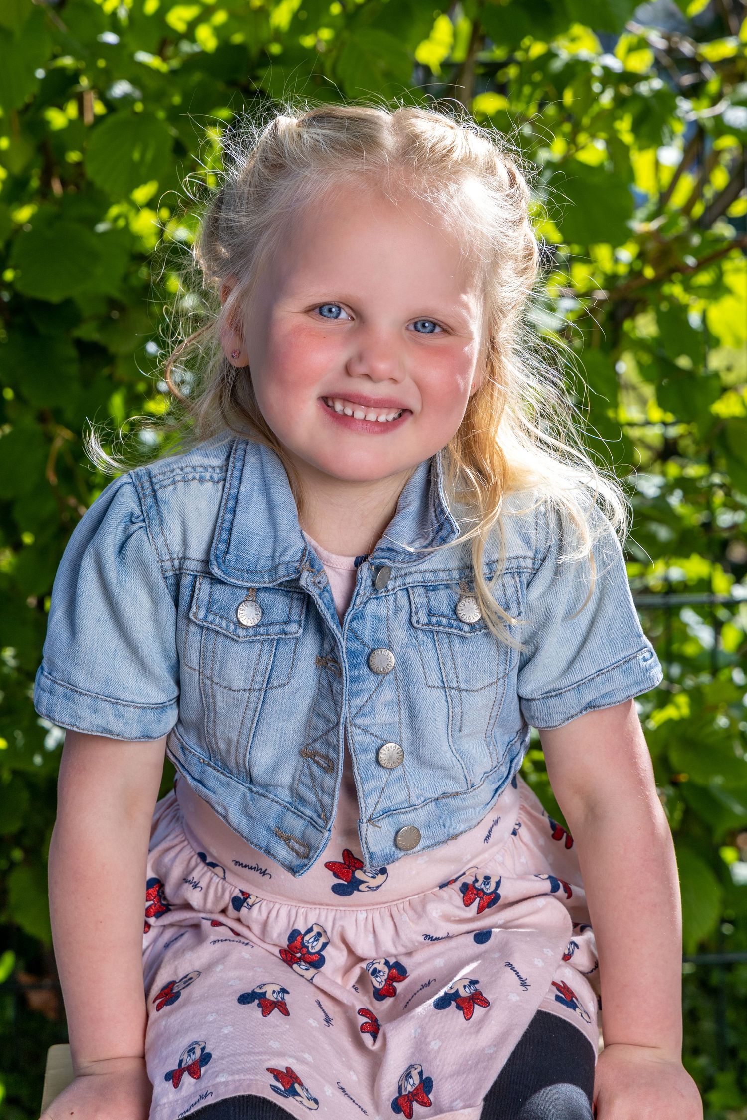 A child in a denim jacket and floral dress sits outdoors against a backdrop of bright green foliage.