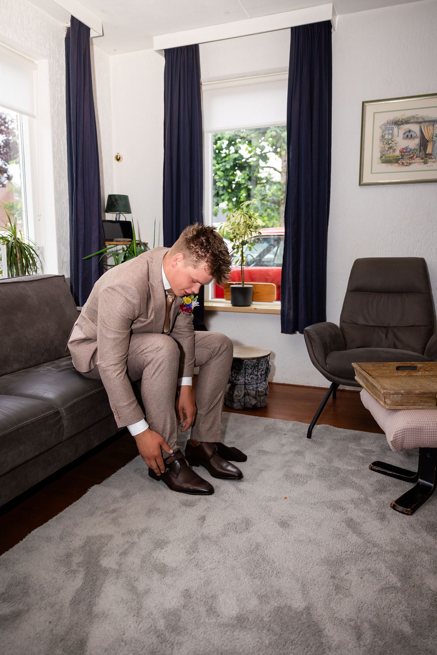 A person in a beige suit adjusting their shoes on grey carpet in a living room.