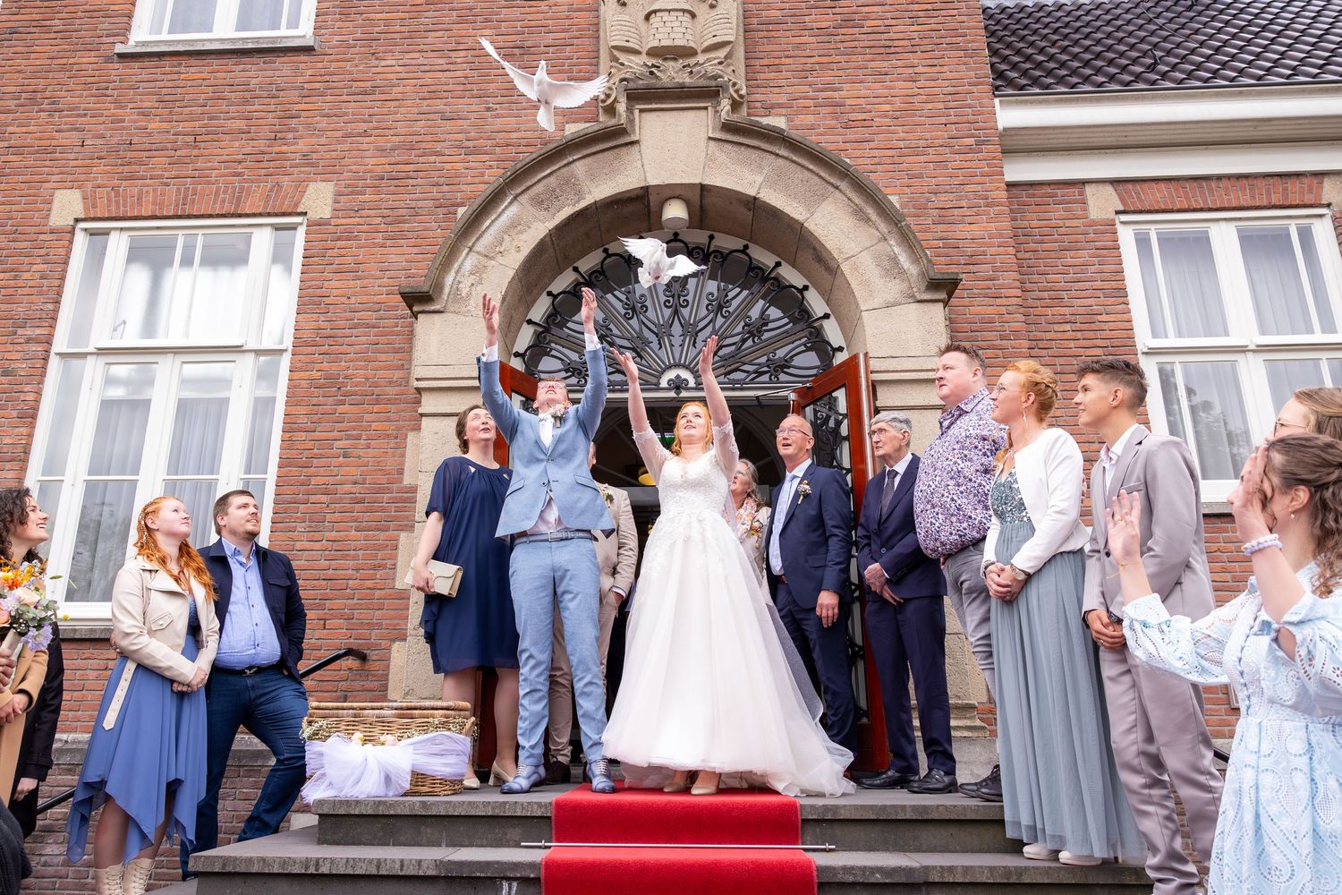 pasgetrouwd stel laat twee witte duiven los en gasten kijken toe voor het gemeentehuis met rode loper
