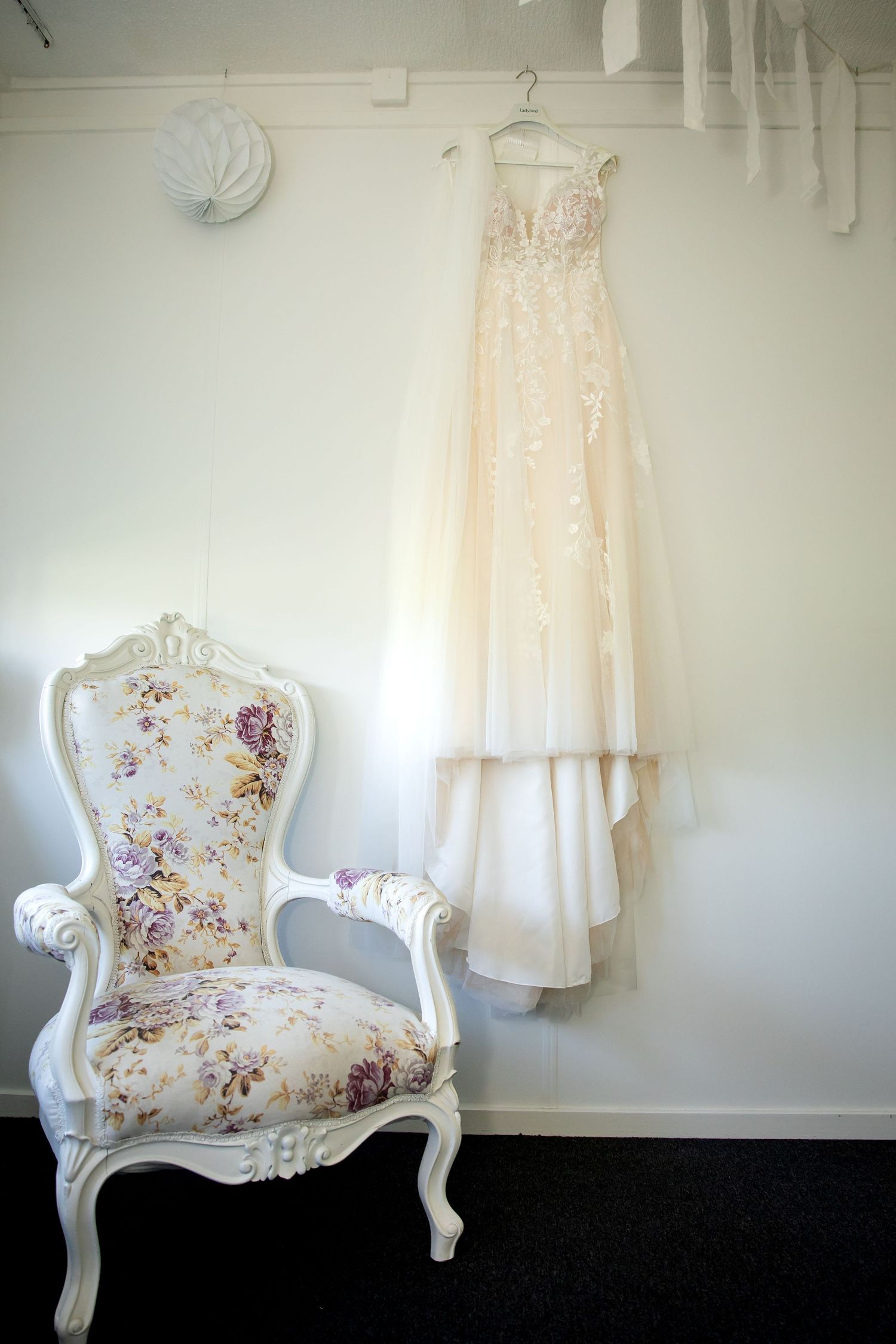 Elegant white floral armchair with cream-colored dress hanging on wall.