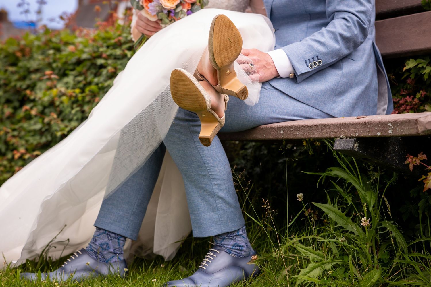 Close-up of wedding dress and suit legs on a wooden bench with grass below.
